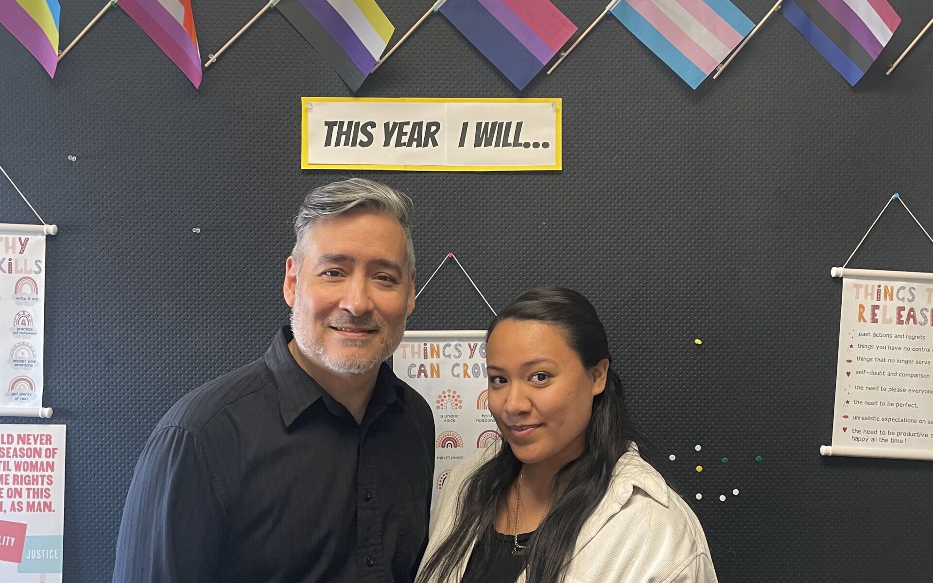 Two teachers pose in front of a black push pin board decorated with Pride flags. On the left is Mr. Sanabria, an older Latine man with light brown skin and short gray hair. He is wearing a long-sleeve black button-up and gray pants. On the right is Ms. Huerta, a young Latine women with light brown skin and long dark brown straight hair. She is wearing a white jacket over a black shirt and black pants. These two teachers run the Pride and Ally Club at ITW David Speer Academy in Chicago, IL.