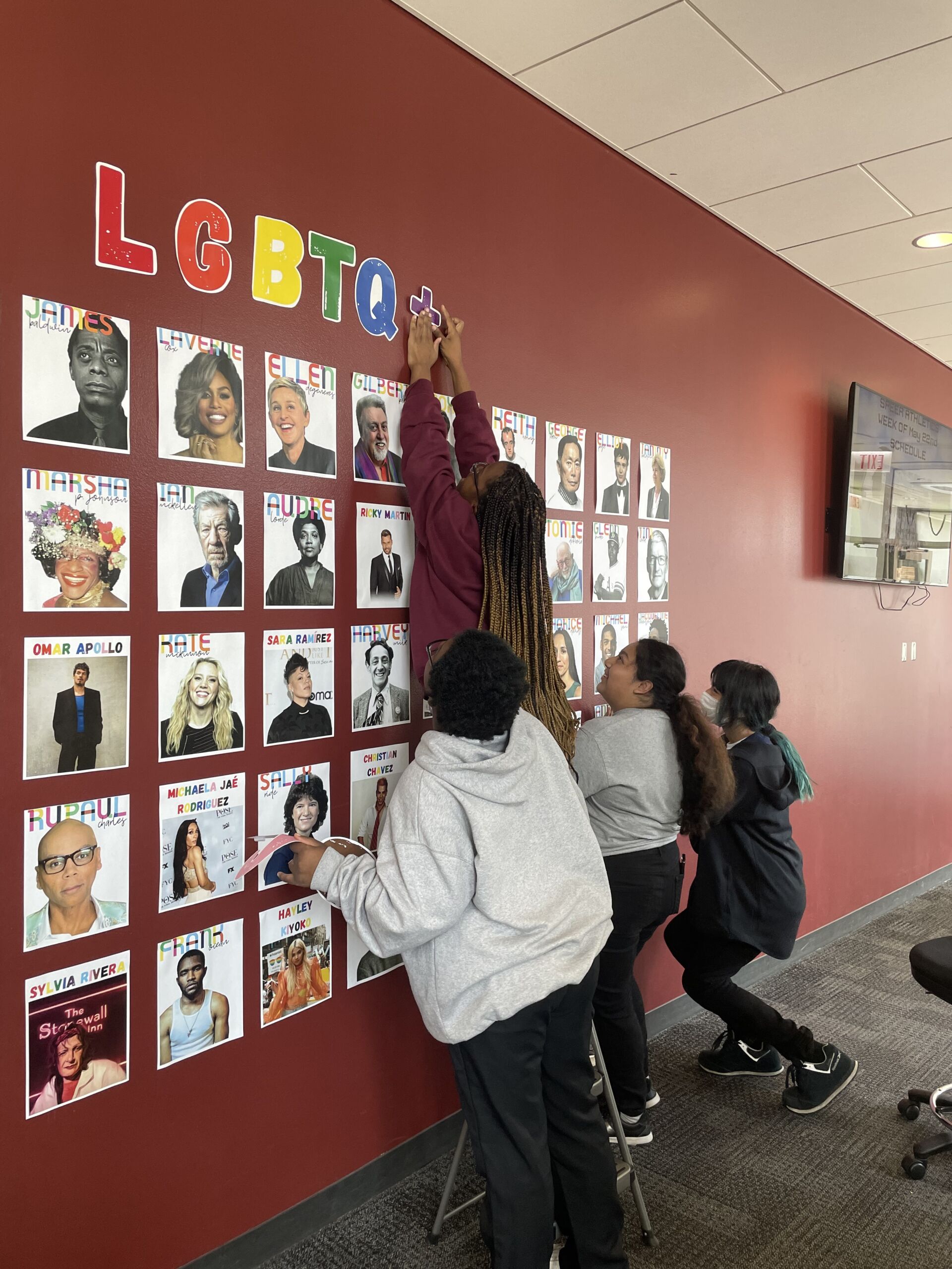 Four students at ITW David Speer Academy work together to put up decorations for Pride Month. One student is standing on a chair and taping up the plus sign in LGBTQ+ on the wall over the top of many photos of famous queer people. The other three students are looking up at them.