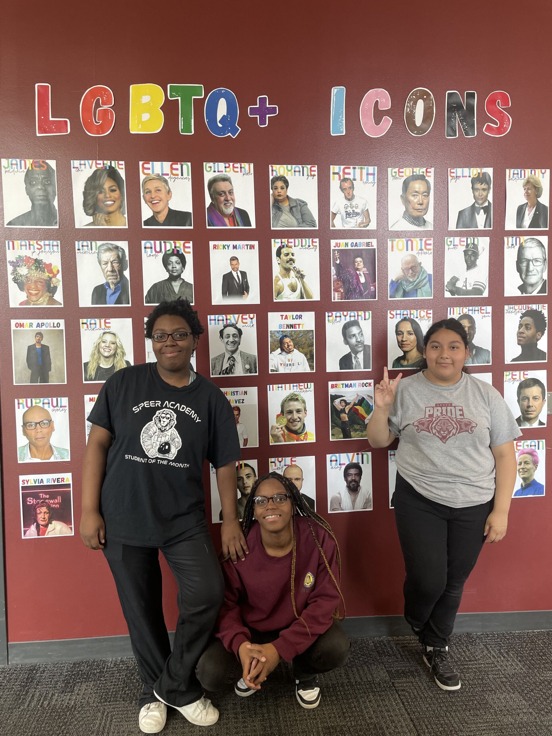 Three students at ITW David Speer Academy pose in front of the finished LGBTQ+ Icons wall decoration. The student standing on the left is a young Black student with short dark brown hair and they are wearing a graphic t-shirt and black pants. To their right, sitting on the ground, is another young Black student with a long sleeved Speer sweatshirt and black pants on. The student standing on the right is a young Latine person with a gray Speer t-shirt and black pants. They are throwing up a peace sign.