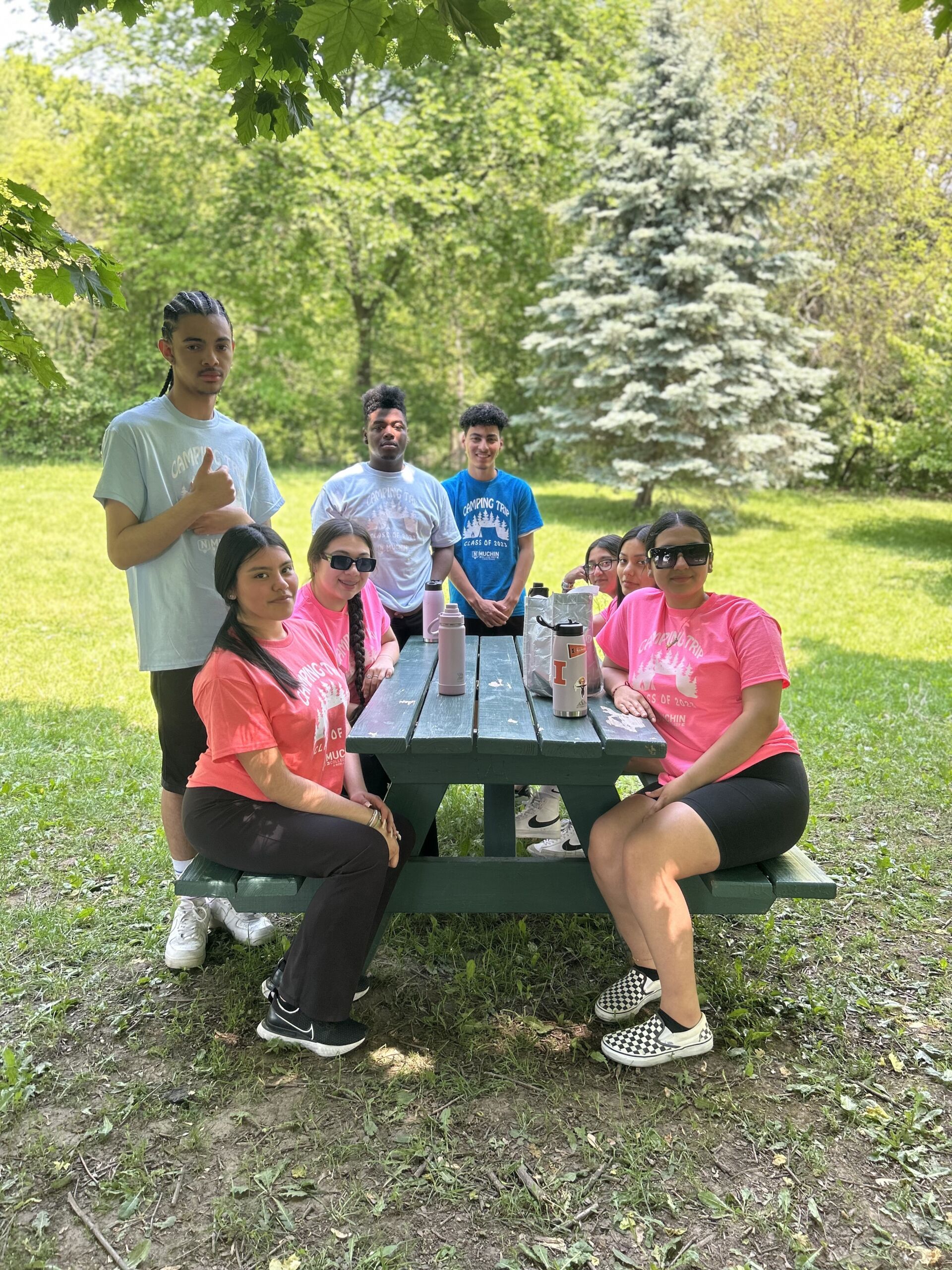 Students are gathered together around a table. Four students are sitting on the table seats while others are standing around the table. One student is posing with thumbs up.