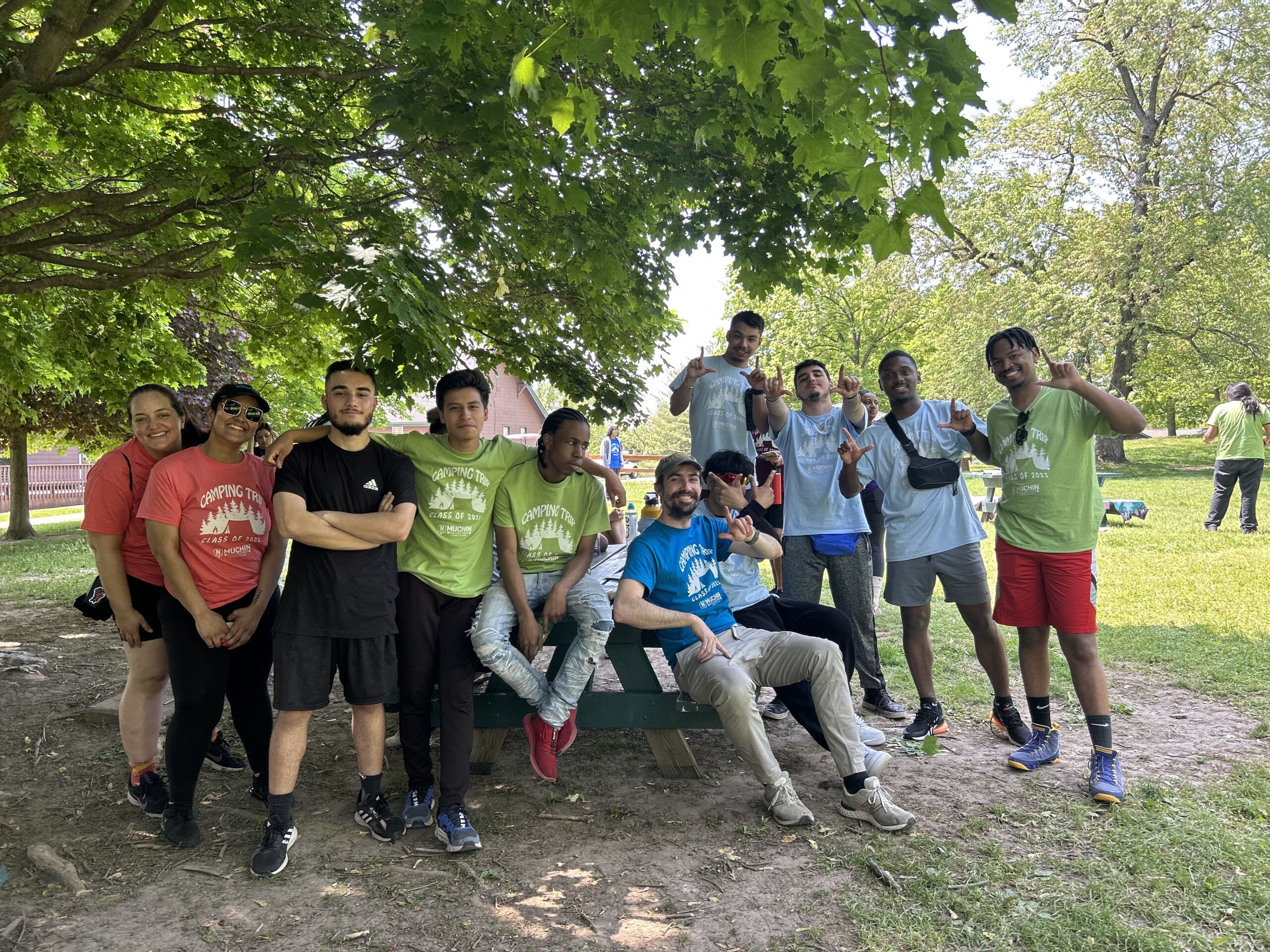 Students pose together on their senior trip. The students are wearing their athletic gear. Students are gathered underneath a tree and one of the teachers is sitting on a chair.