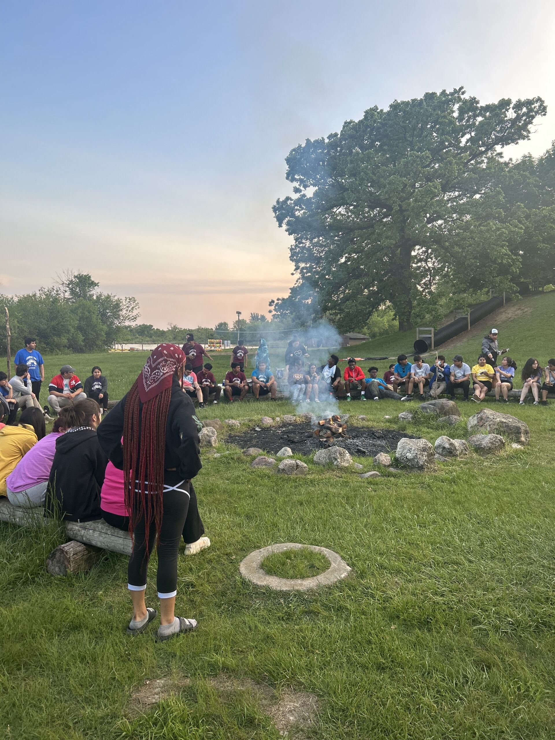 Students gather around the campfire during their senior trip. Students are sitting or standing up. Some are on their phones and others are talking to each other. The campfire has smoke coming out.