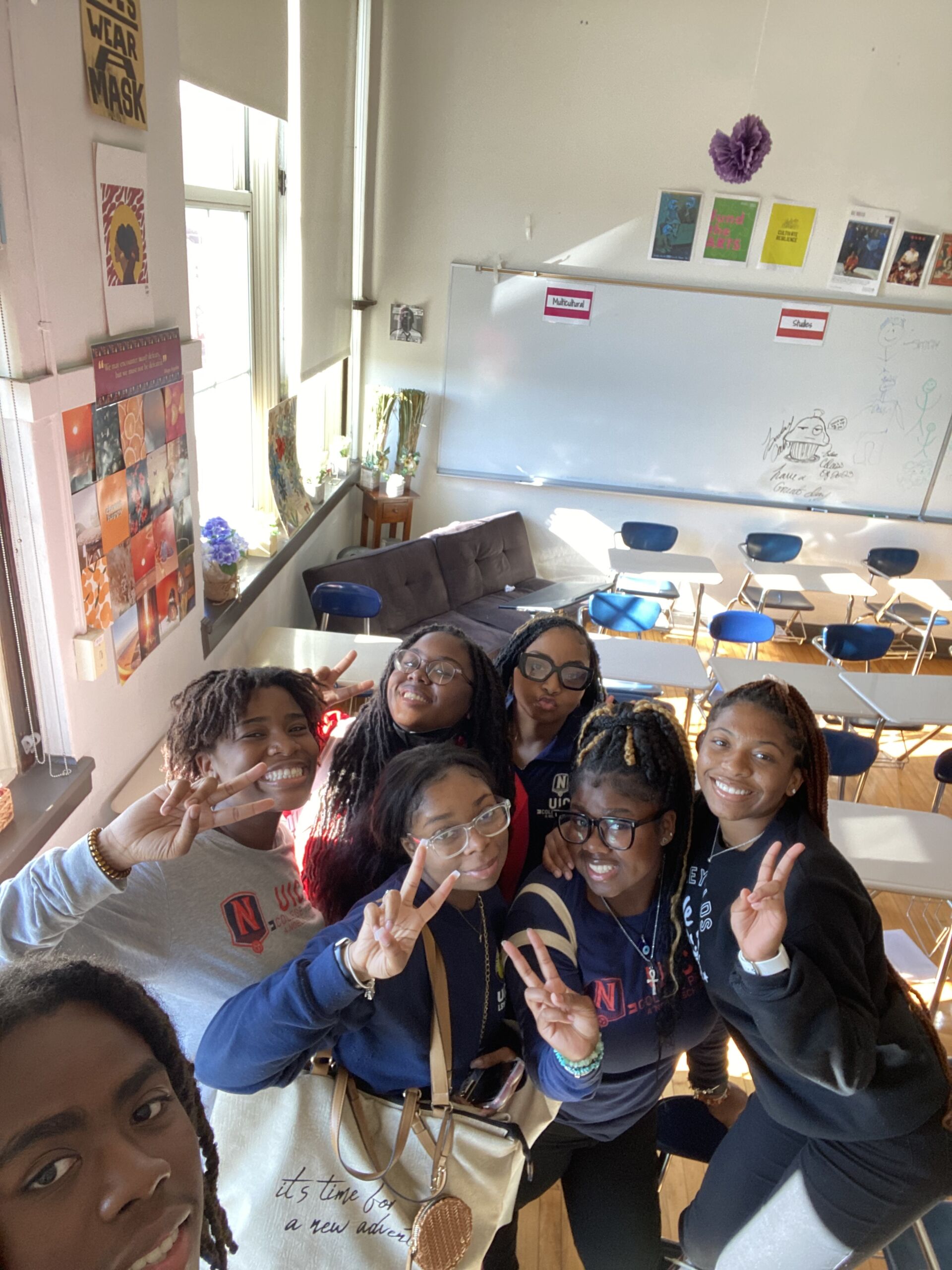 Seven students are pictured in the selfie above. Students are smiling and posing. Some students are sticking out the peace sign. Four students in the middle are wearing glasses. The students took the photo inside the classroom.