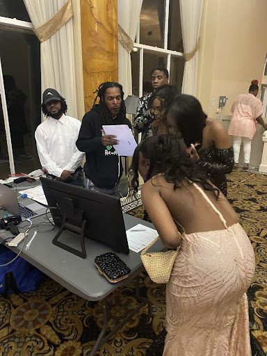 Students gather around the photographers laptop as he shows them the photos he took. Students are wearing dresses and suits. One student, who's face is not visible, is wearing a coral, light pink dress. Another student is wearing a white button up long sleeve dress shirt. Another student is wearing a sparkly black dress.