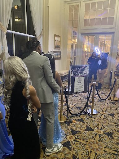 Students wait in line to get a 360 photo of themselves. Students in the front of the line, faces not visible, are wearing a gray tan suit and a tiffany blue dress. The student behind them is wearing a black dress. In front of the students is the camera set up with lights, camera, and business info on a stand.