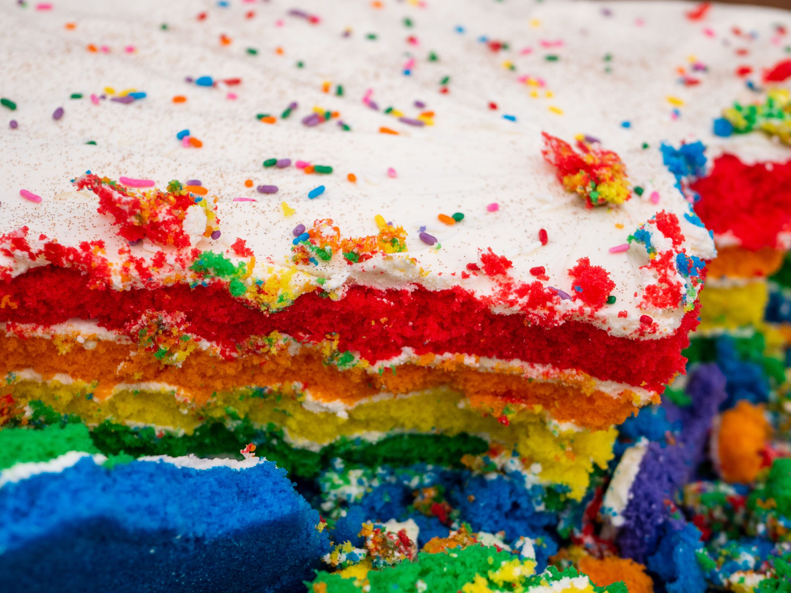 Photo of a rainbow cake. The cake has red, orange, yellow, green, blue, and purple coloring inside the cake. The top of the cake has white icing with rainbow sprinkles.
