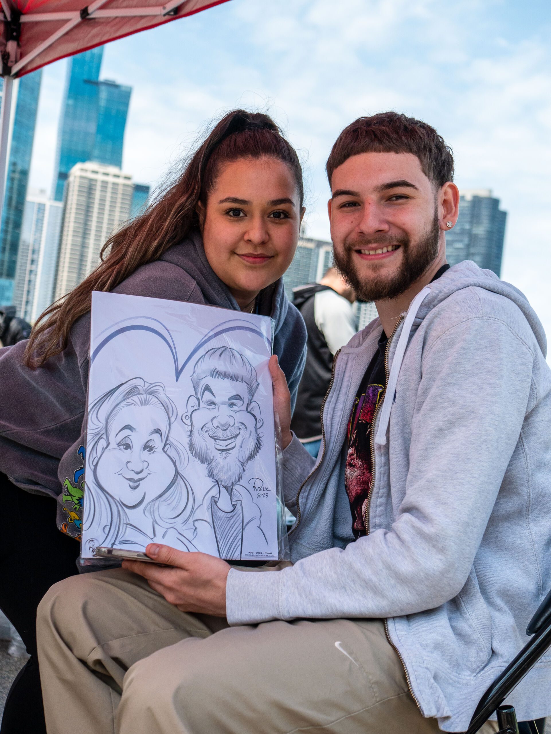Two people pose as they show off their drawing. The two received a drawing from a carnival vendor. The person on the left has her hair up on a ponytail and the person on the right is wearing a gray sweater. He is sitting down on a chair.