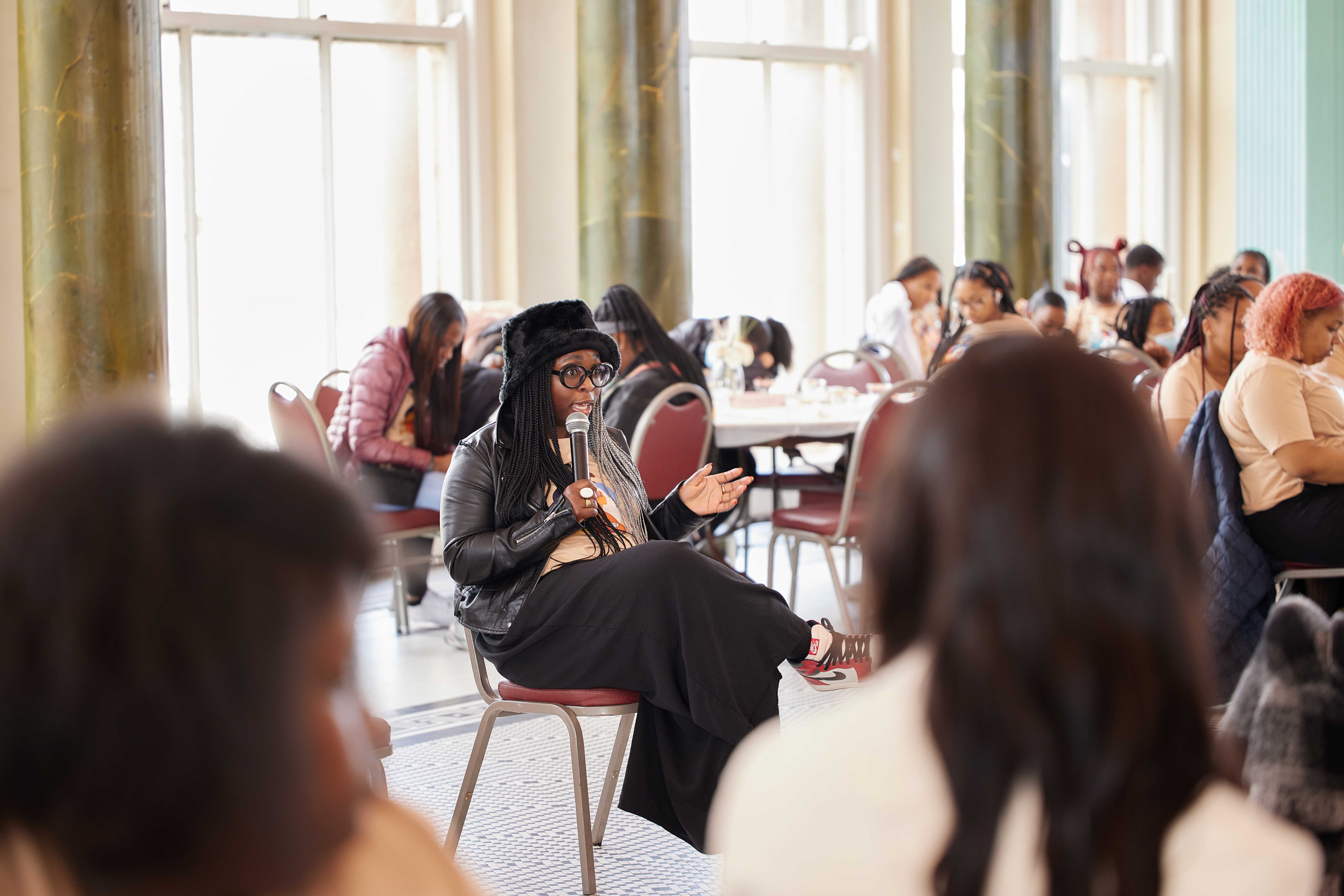 Photo shows IB Majekodunmi, a successful entrepreneur and the founder of Refine Collective, sitting in a chair and holding a microphone, addressing the crowd at the Johnson College Prep Women's Empowerment Brunch. She is a young Black woman with long black braids and wearing a fuzzy black bucket hat, a black leather jacket over the beige JCP Women's Empowerment Brunch graphic tee, loose black dress pants, and black, red, and white Nike Air sneakers. She is talking with her hands and her mouth and eyes are open wide as she speaks. She is surrounded in the background and foreground of the image by the audience.