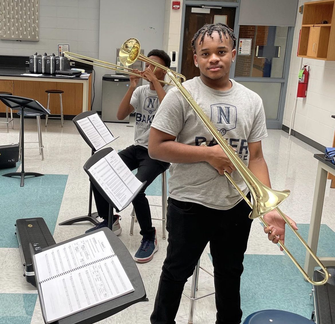 A student is standing holding their trumpet in their jazz class. The student is wearing a Baker graphic tee and black pants. The student is holding the trumpet with his right hand and left. The student is inside an orchestra/music room with music sheets on the stands. There is another student behind the first student but their face is not visible.