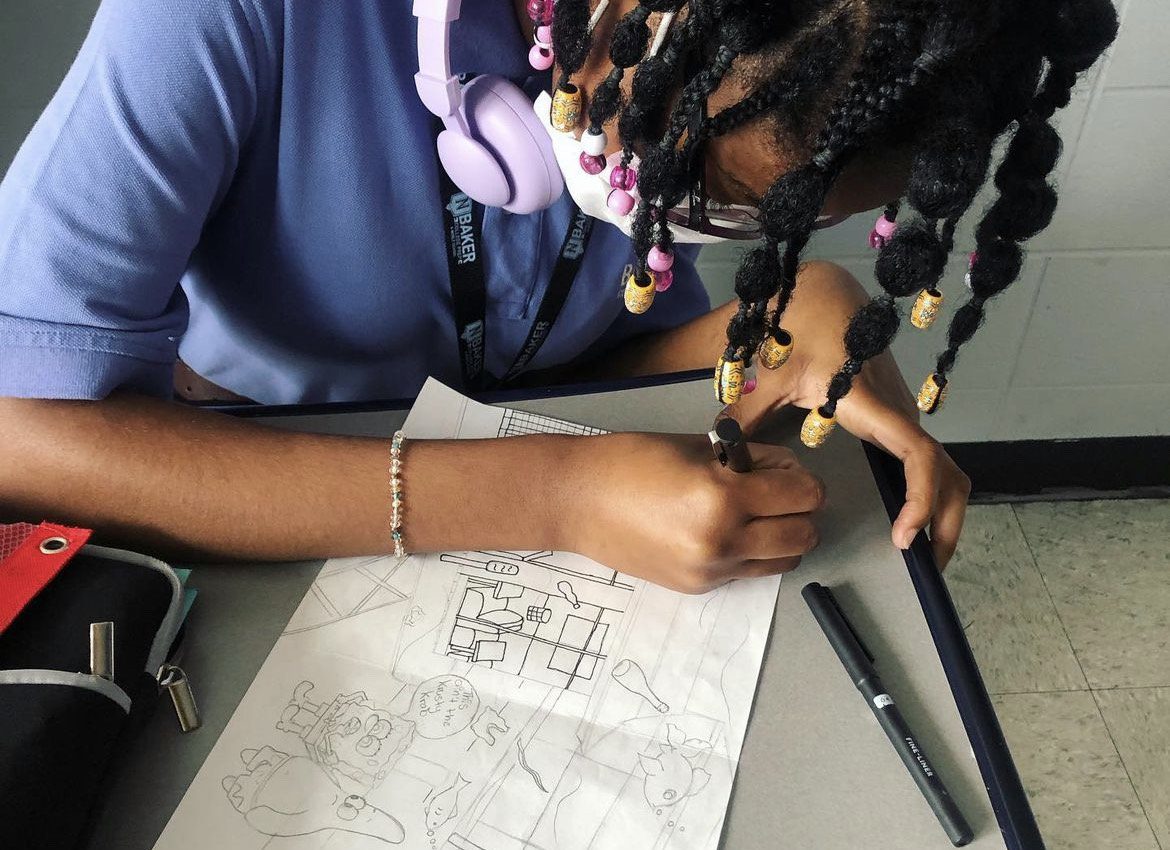 A sophomore student is pictured drawing in their SkyArt class. The students face is not visible but the student has pink headphone over their neck. The student also has a grayish-blue school shirt on. The student is currently tracing their pencil drawing with a artist pen. The drawing looks to be a scene of Spongebob Squarepants.