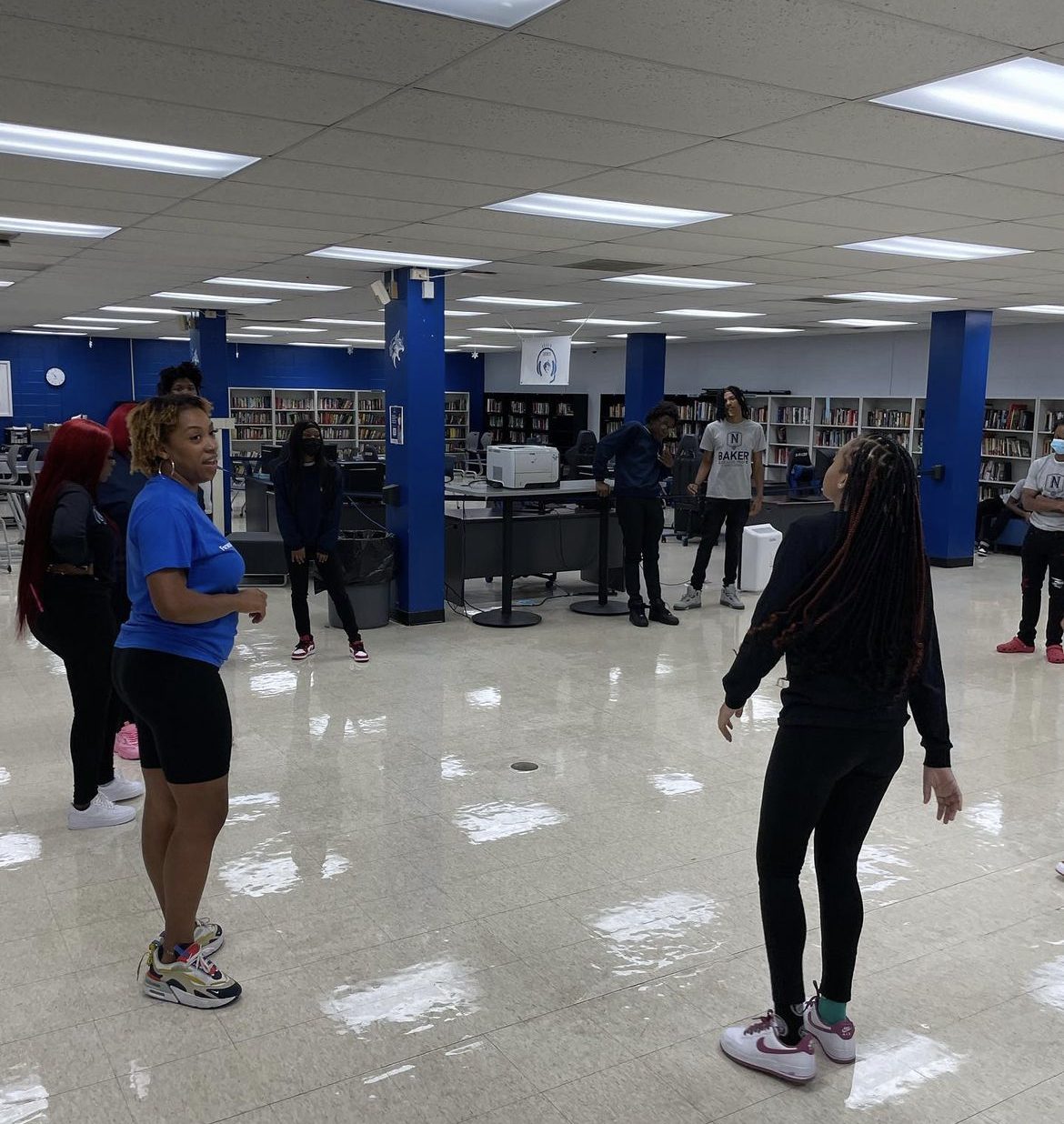Students are spread out as they learn new choreography from their dance instructor. The students are wearing comfortable clothing. Students who are visible are wearing Baker graphic tees and jogging pants. The instructor leads the class as she wears a bright blue shirt and black shorts.