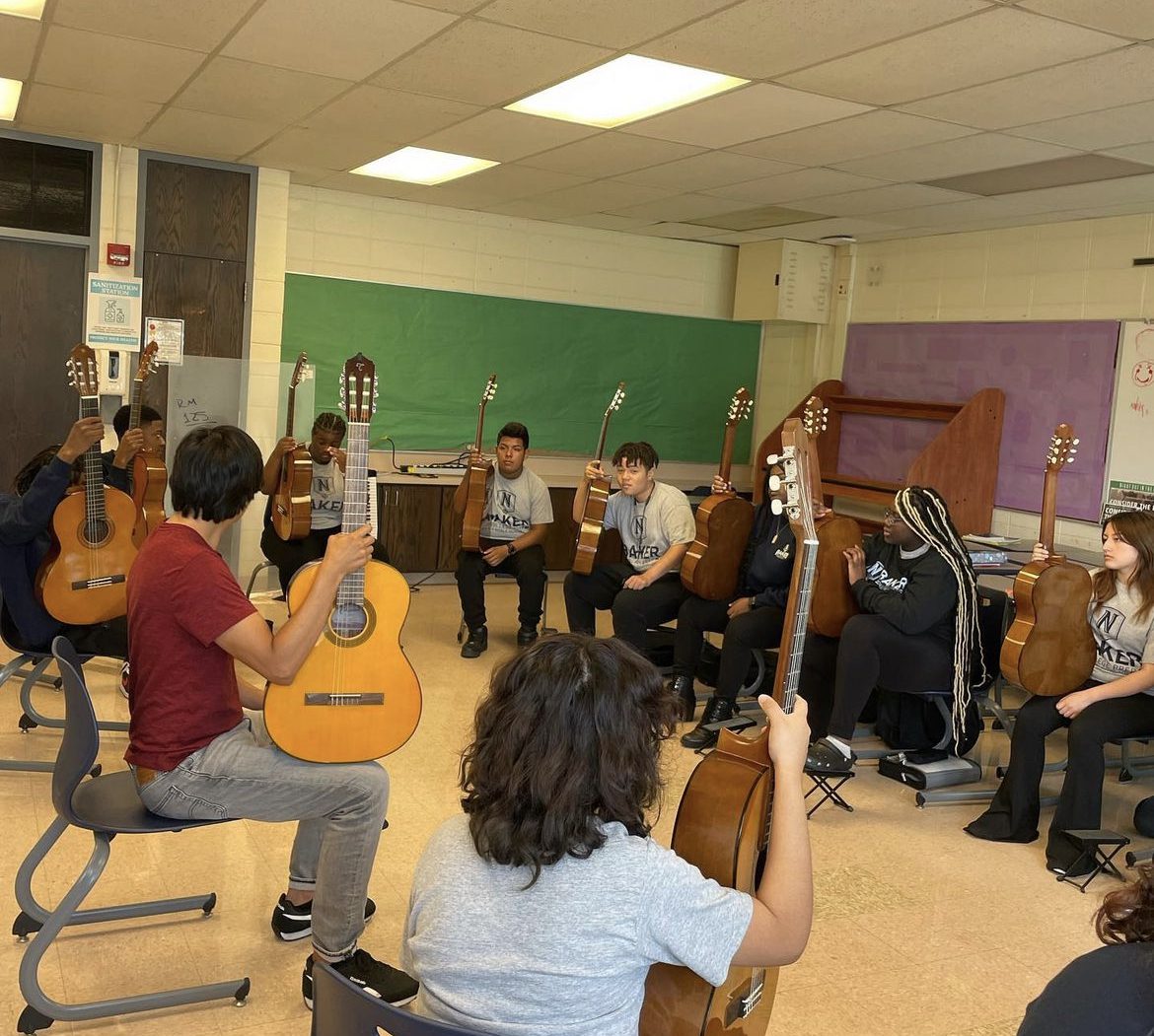 Students are sat in their chairs in a circle. The students are wearing their Baker school graphic tee. The instructor is wearing a read shirt and gray dress pants. The students and instructor are holding their guitar upright on top of their knees.