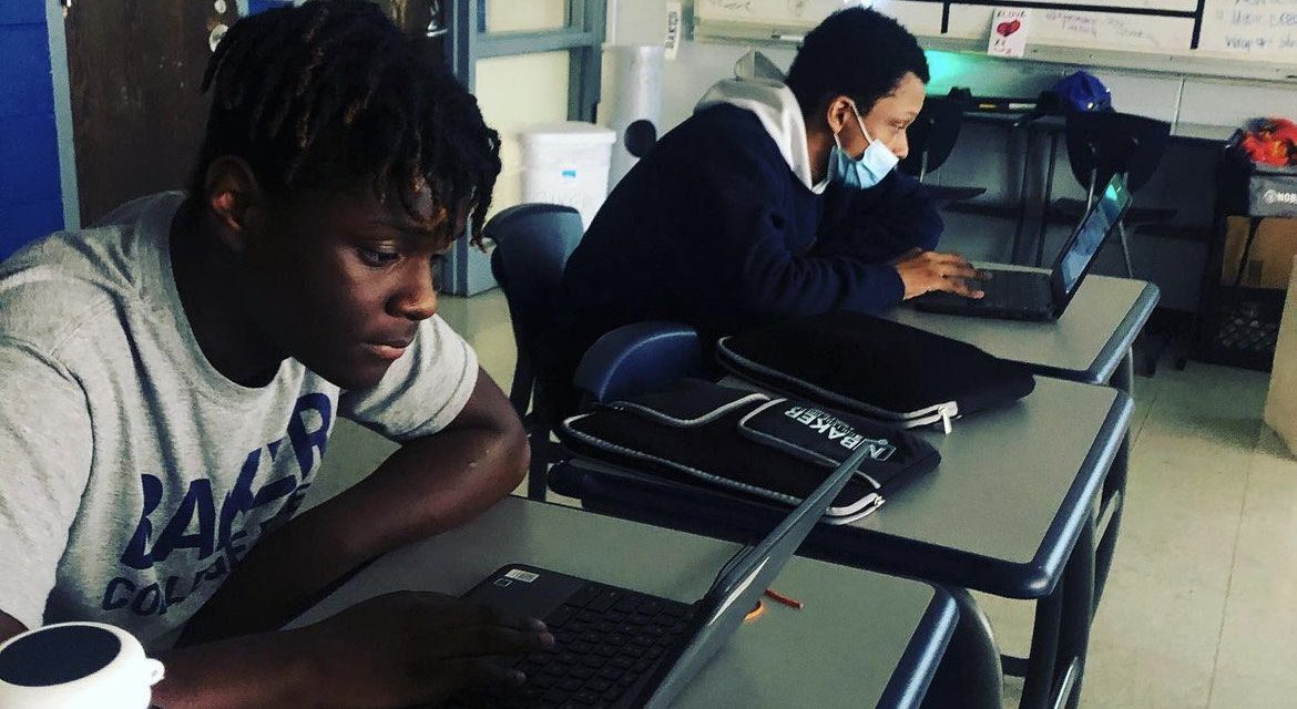 Two students are visible working on their computer in their Computer Science-Project Stem-Amazon Future Engineer course. The students are wearing their Baker uniform. One student is wearing a mask on his face. The other student, closest to the camera, is not. The two are both sitting one desk apart in the classroom.