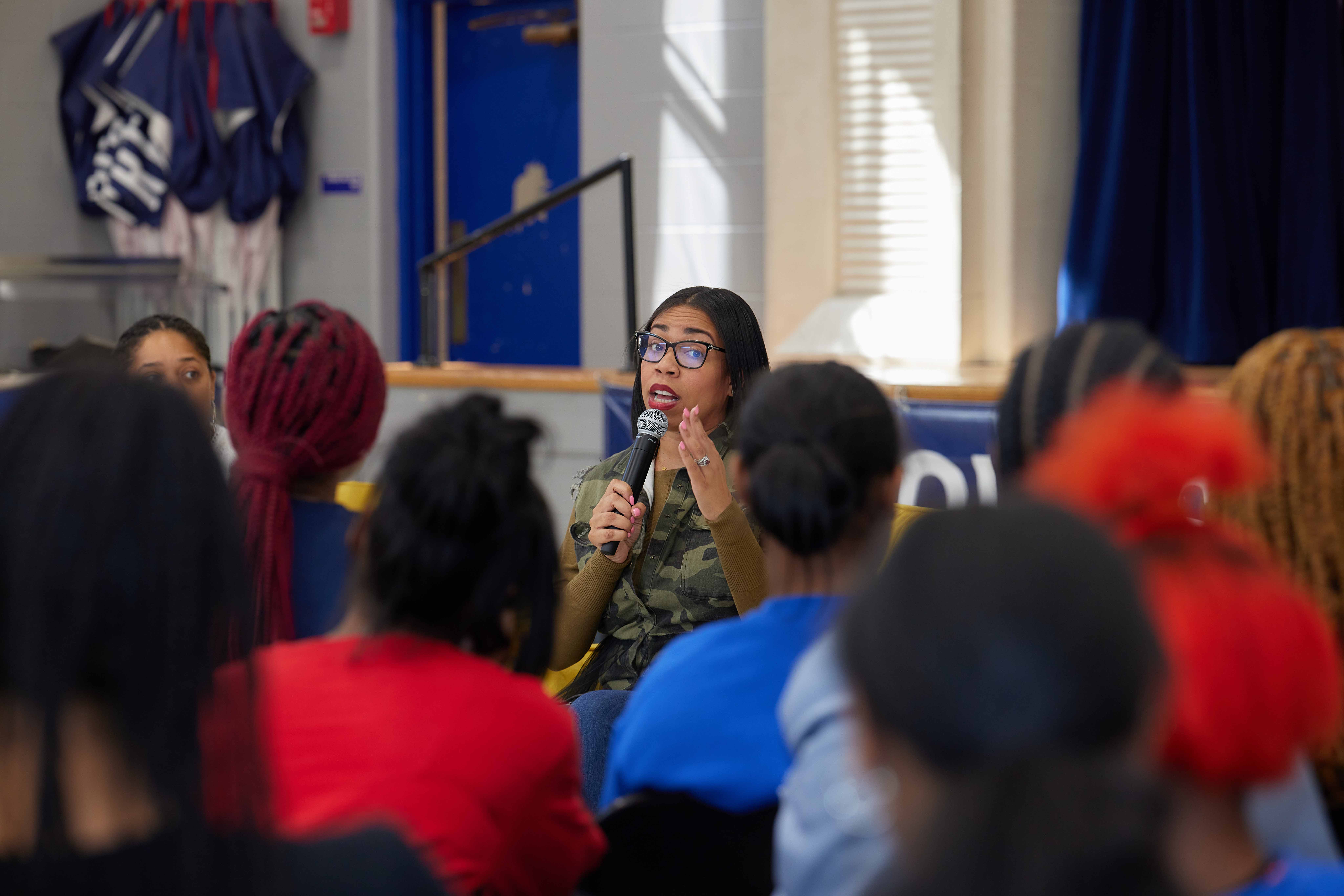 Image shows Jennifer Reid Davis, Noble Schools' Head of Strategy and Equity, speaking in front of female students at Johnson College Prep in Chicago, Illinois. Jennifer is sitting down and holding a mic in her hand. Her face is animated and looks like she is in the middle of telling a great story. In the foreground of the image is the blurred out heads and shoulders of the audience, framing Jennifer in the center of the photo.
