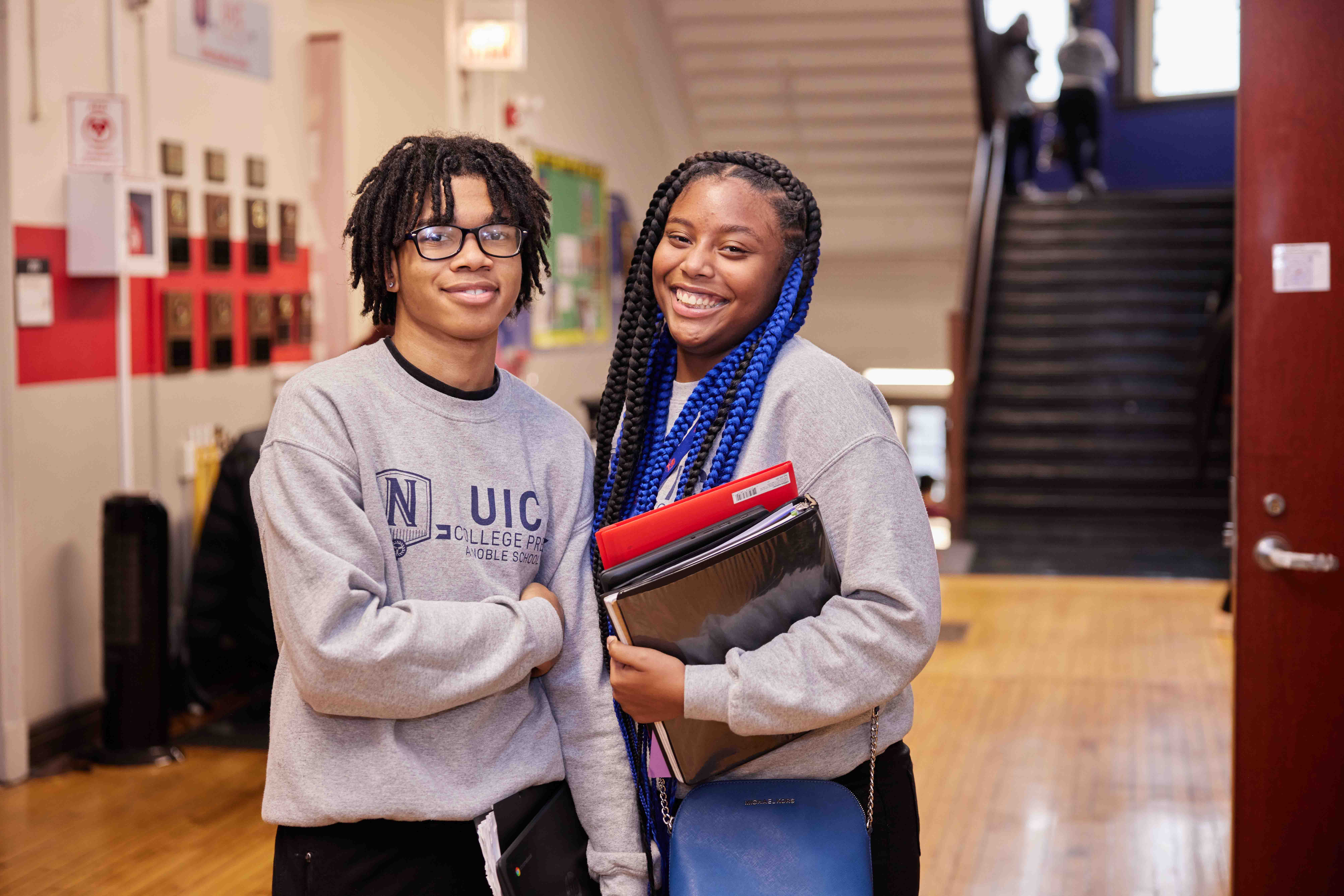 Photo shows two students at UIC College Prep in Chicago, Illinois standing in the hallway of the school building. The student on the left is a young Black man with medium-length locs. He is wearing a light grey sweatshirt with the dark blue UIC College Prep logo on it. The student on the right is a young Black girl. She has long black and blue braids. She is wearing the same light grey sweatshirt and holding a binder, a folder, and a laptop with one arm. They are leaning into each other and smiling at the camera.