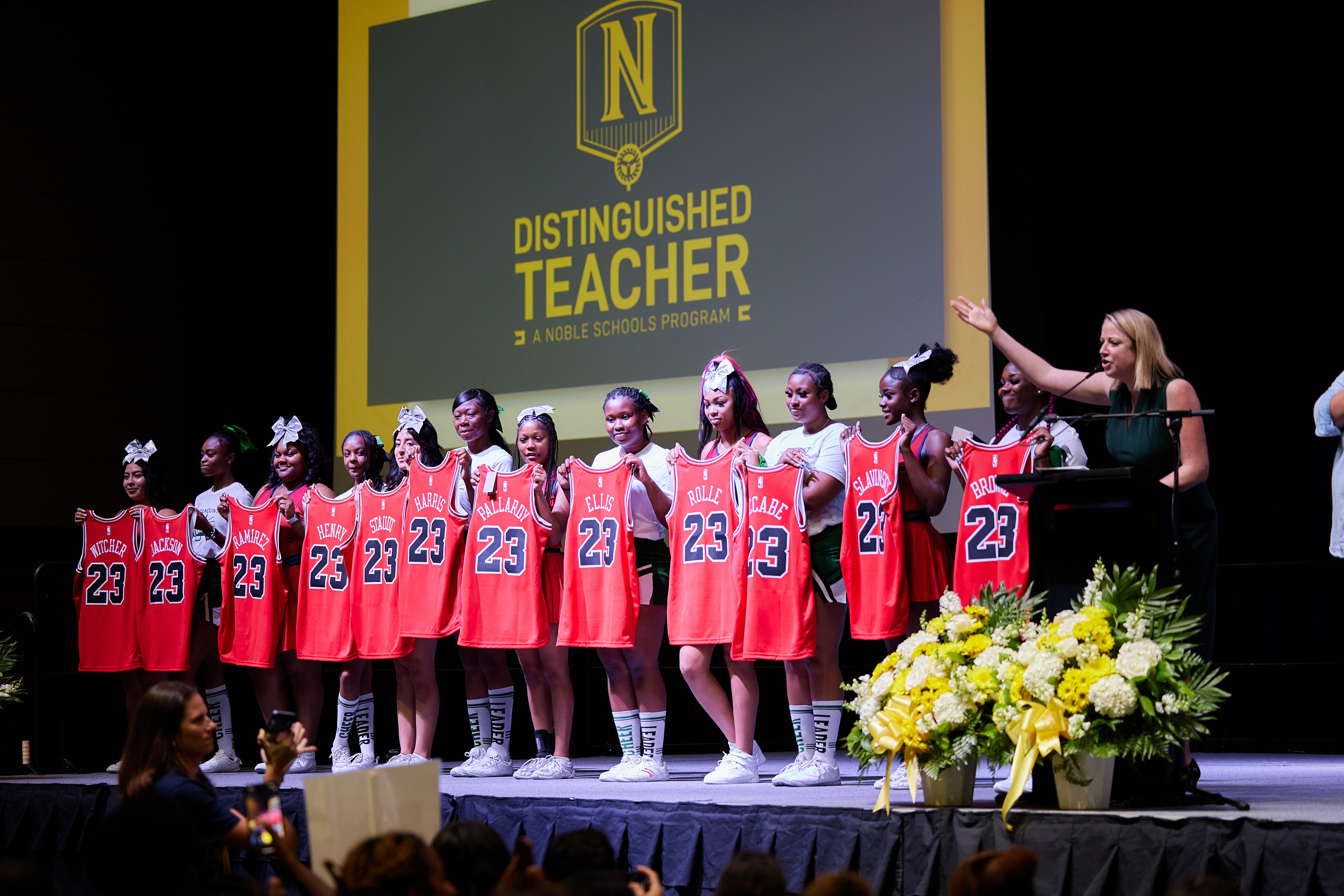 Photo shows the Chicago Bulls College Prep cheer team on the stage presenting personalized jerseys for the Noble Schools' Distinguished Teacher 2023 cohort. To their right is Ellen Metz, Head of Schools at Noble Schools, who is at the podium and speaking to the crowd, her hand raised towards the jerseys to present them. Behind them is a projector screen that shows the Noble Distinguished Teacher Program logo.
