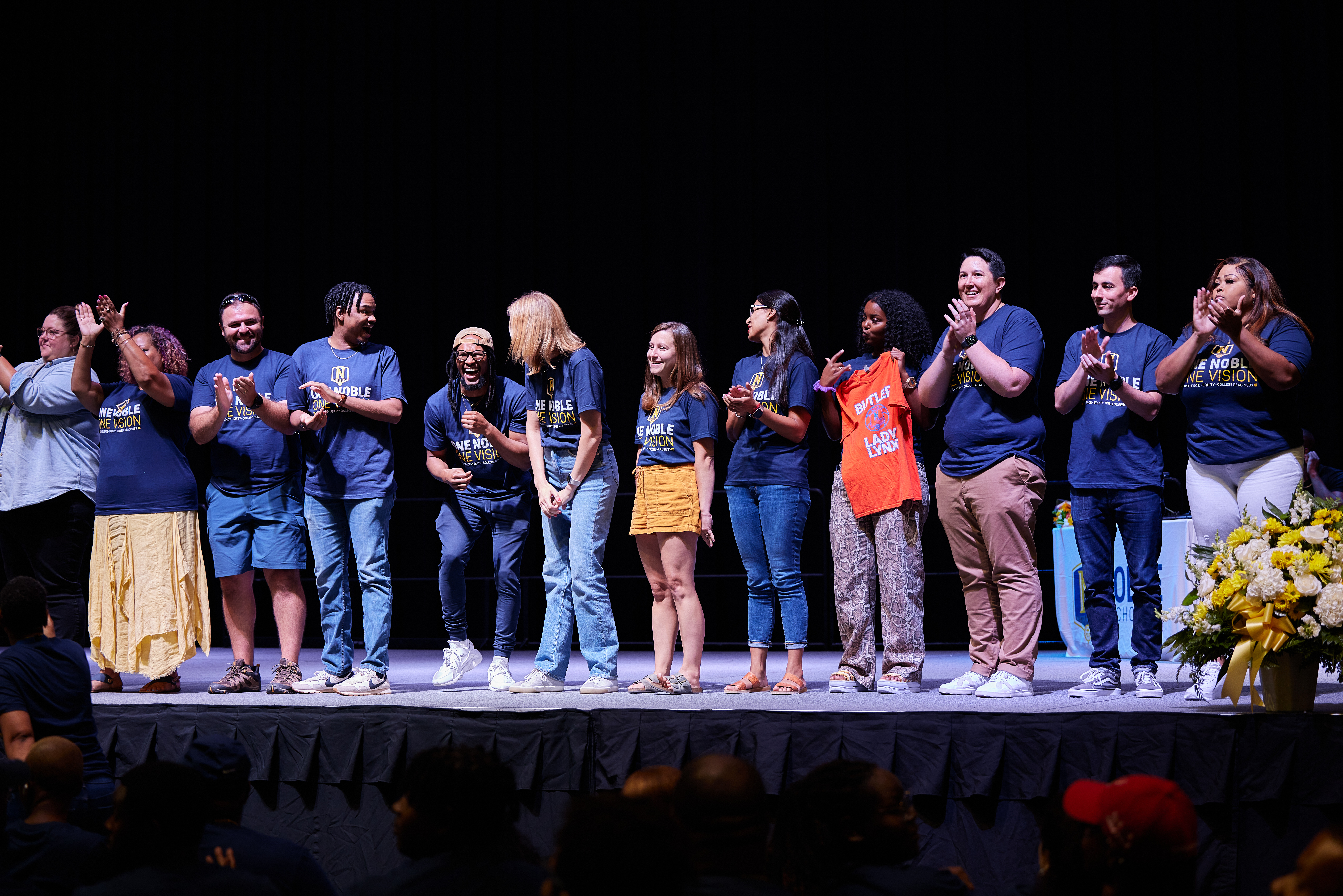 Photo shows a wide shot of the entire Noble Schools' Distinguished Teacher 2023 cohort lined up on stage. They are all smiling and clapping as they are surprised to be honored at the kickoff event.