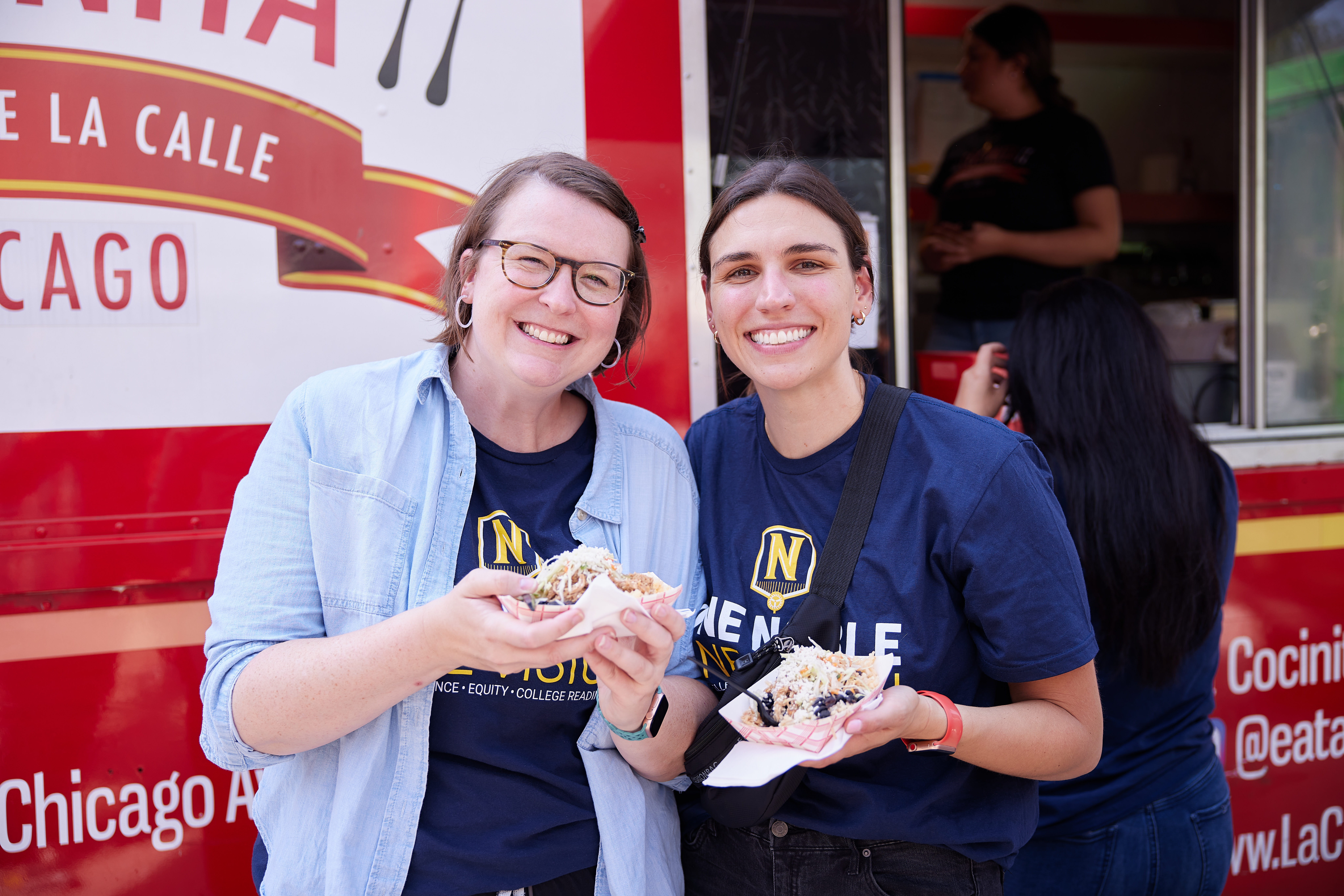 Photo shows two Noble Schools staff members holding food in front of the La Concina food truck. They are smiling and looking at the camera.