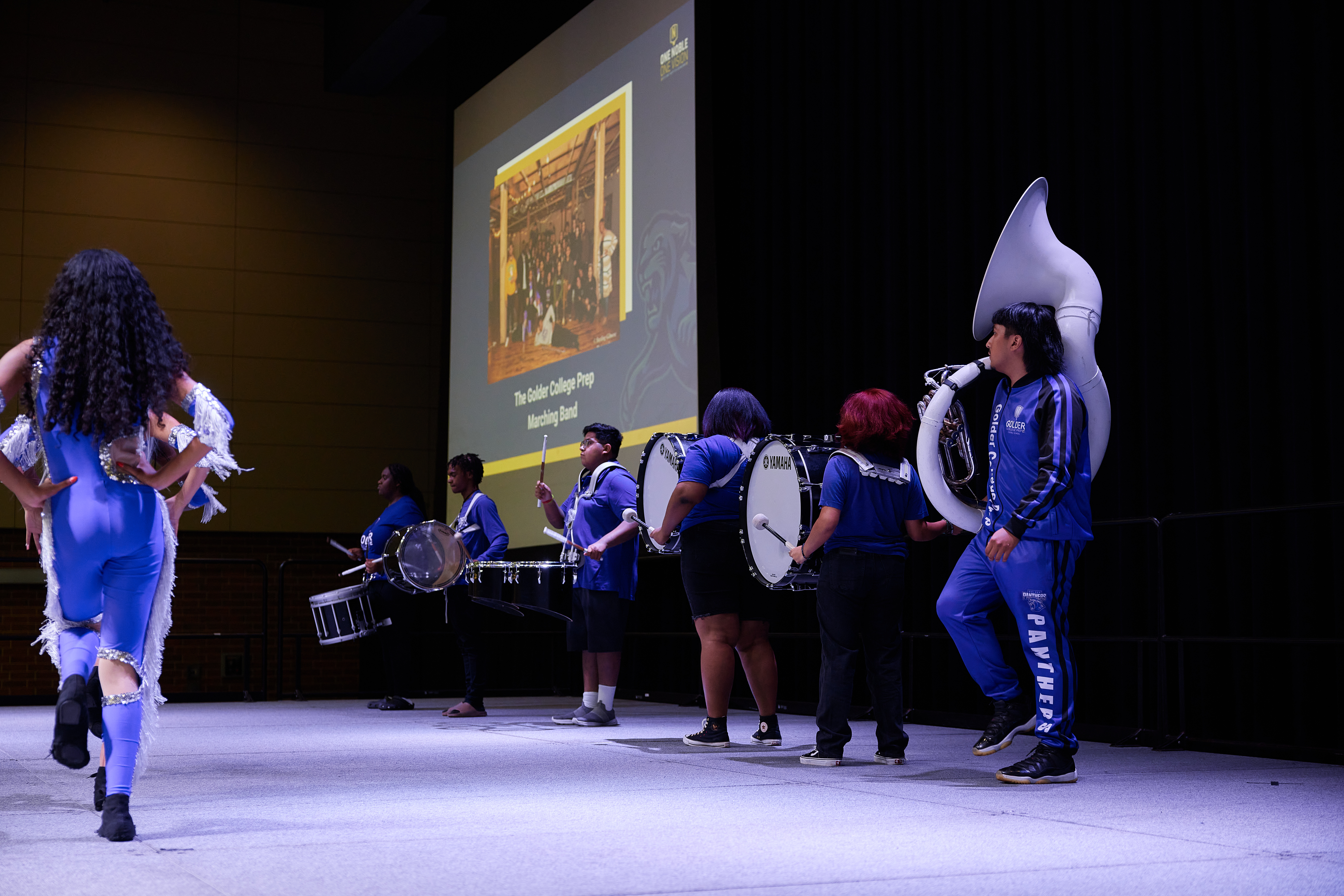 Photo shows the Golder College Prep marching band standing on stage, performing at the Noble School's Staff Kickoff for the 2023-2024 school year. The dancers of the band dance are strutting in the front as a tuba player and multiple drum players play behind them. There is a big projector screen behind them that says "The Golder College Prep Marching Band".
