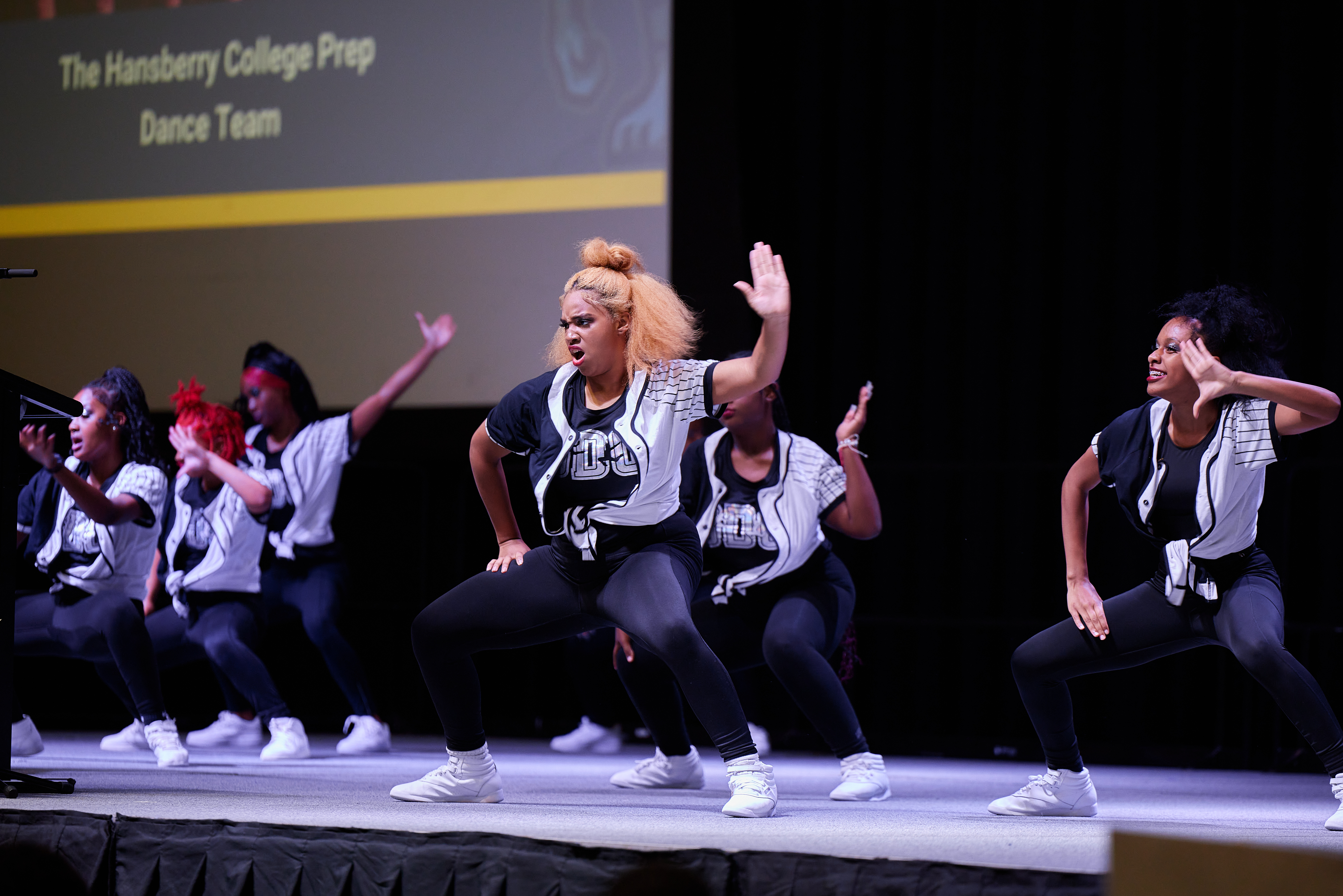 Photo shows a wide shot of the Hansberry dance team, the Bengal Dance Collective, performing on stage at the Noble Schools' Staff Kickoff for the 2023-2024 school year. They are all bent low at the knees with their right hand on their thigh and their left hand raised in the air. The dancer in the center of the photo has a deep look of concentration and effort on her face.
