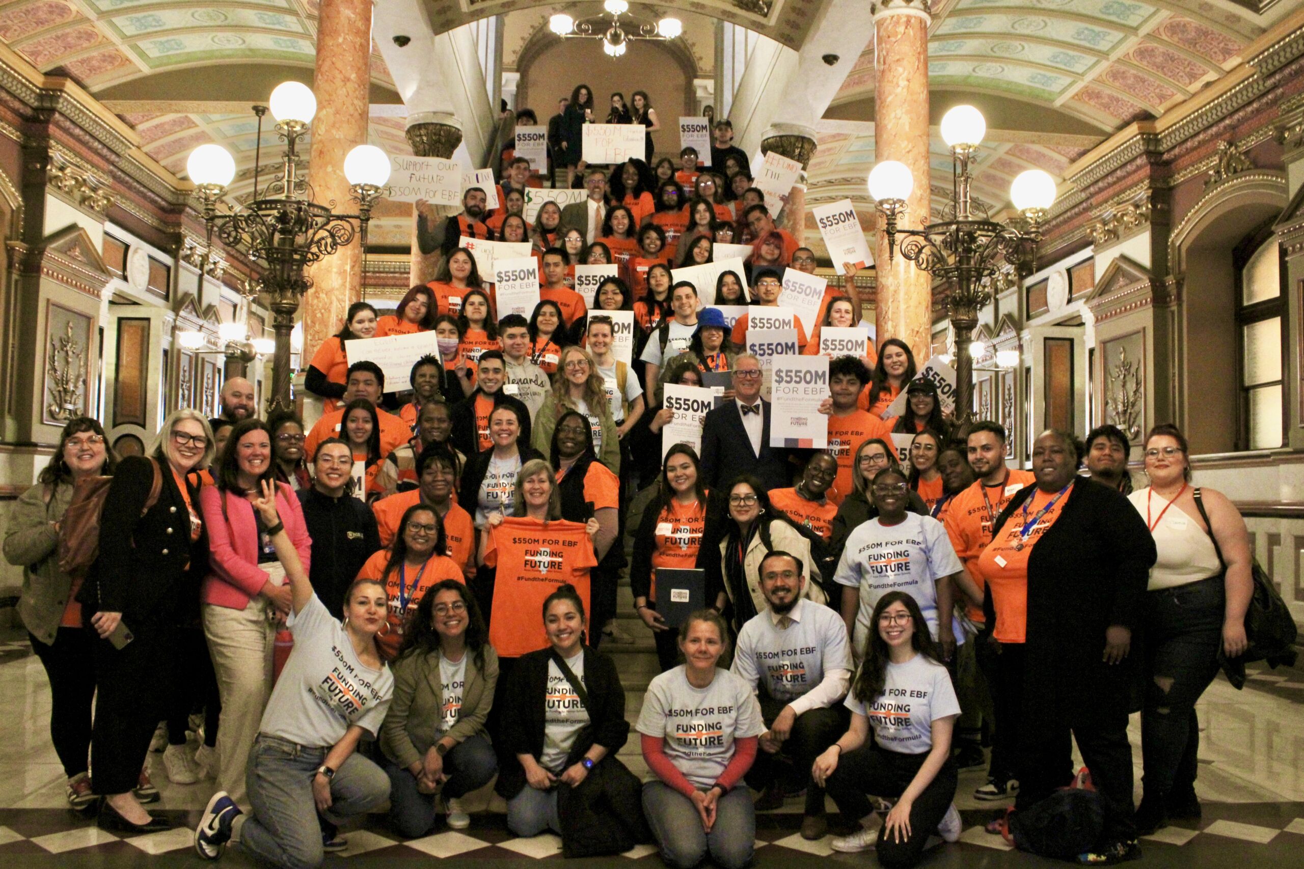 Photo shows a group shot of everyone who showed up in April 2023 to advocate for evidence-based funding for Illinois schools. The group is standing and posing up the stairs of the Illinois State Capitol building. This group includes Noble Schools' parents and staff, the team at Funding Illinois Future, and other Illinois public schools parents, staff, and community members. Most people are smiling and wearing bright orange shirts that read "$550M for Evidence-Based Funding" and have the Funding Illinois Future logo on them. Many people in the back and at the top of the stairs are holding signs that say the same thing.