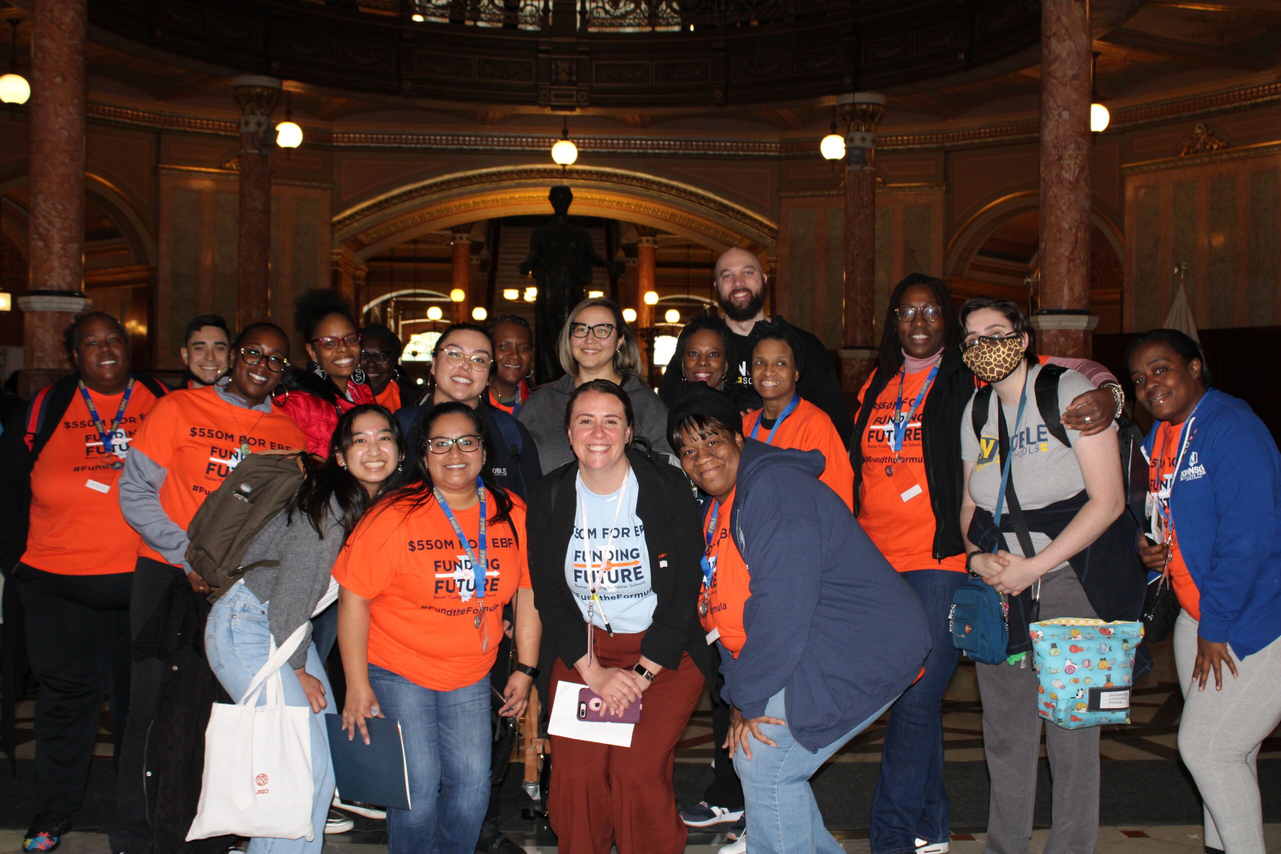 Image shows a group photo of Noble Schools' parents and staff standing in the Illinois State Capitol building's rotunda. Everyone is smiling and many are wearing bright orange shirts that say "$550M for Evidence-Based Funding" with the Funding Illinois Future logo on them. This photo was taken during Noble Schools' Lobby Day trip in April 2023.