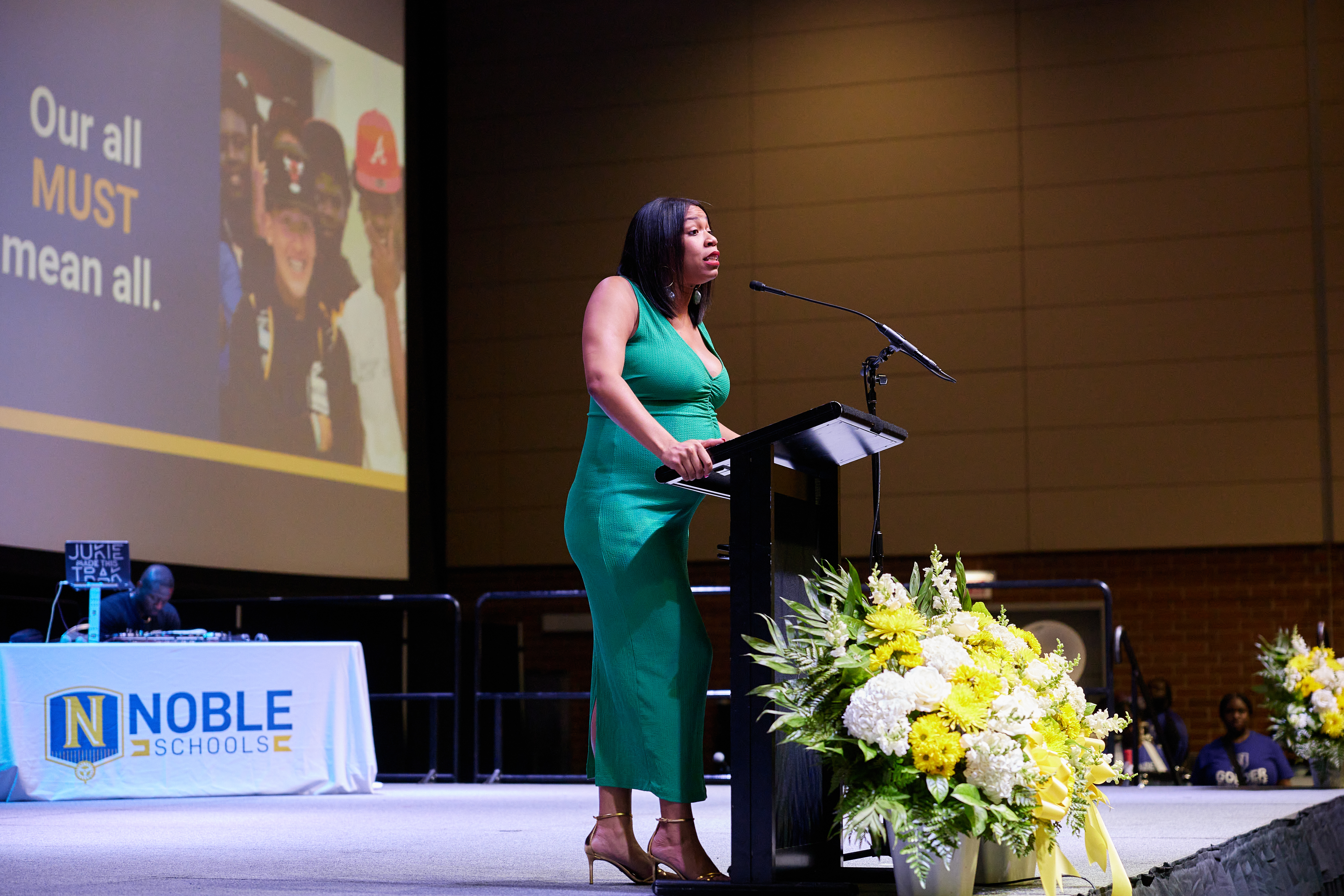 Photo shows a full body shot of Jennifer Reid Davis, the Head of Equity and Strategy at Noble Schools, as she addresses the crowd on stage at the podium. Behind her is a projector screen that has a photo of Chicago Bulls College Prep students next to words that read "Our all MUST mean all".