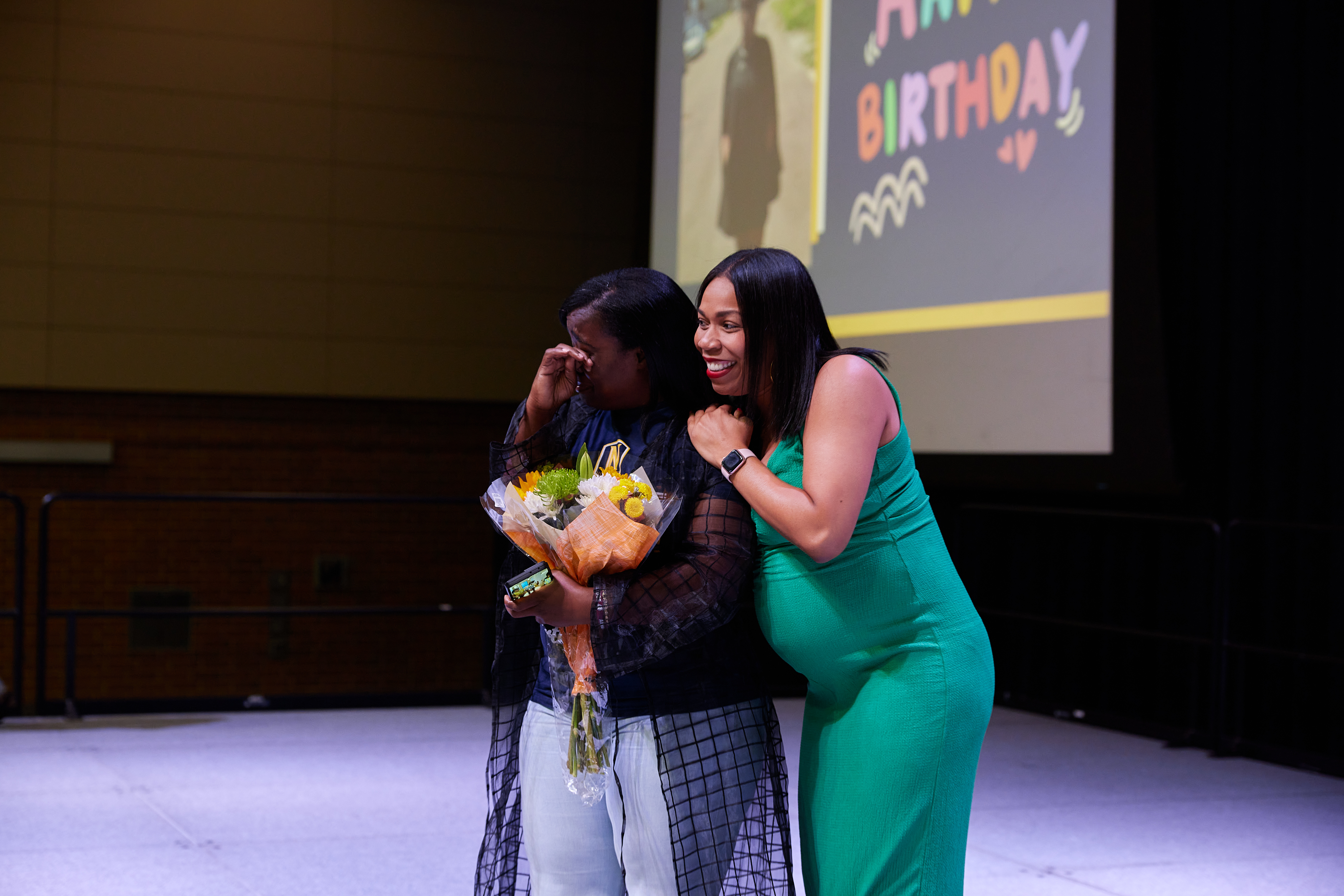 In this photo, Noble Schools' parent Myisha Shields is on stage, side hugging Jennifer Reid Davis, Noble's Head of Strategy and Equity. Ms Shields is holding a bouquet of flowers and crying. Behind both of them is a projector screen that has a photo of Ms Shield's on it and the words "Happy Birthday".