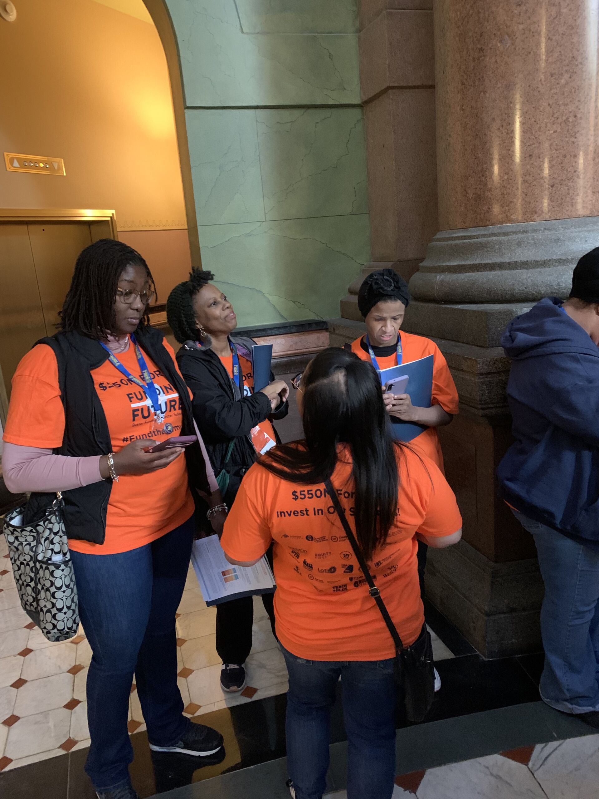 Image shows Noble Schools' parents and a staff member talking while they wait to speak with representatives in the hallways of the Illinois State Capitol building. They are all wearing bright orange shirts that read "$550M for Evidence-Based Funding" and have the Illinois Funding Future logo on them. They are holding folders with all the information they need to prep for their talks with representatives.