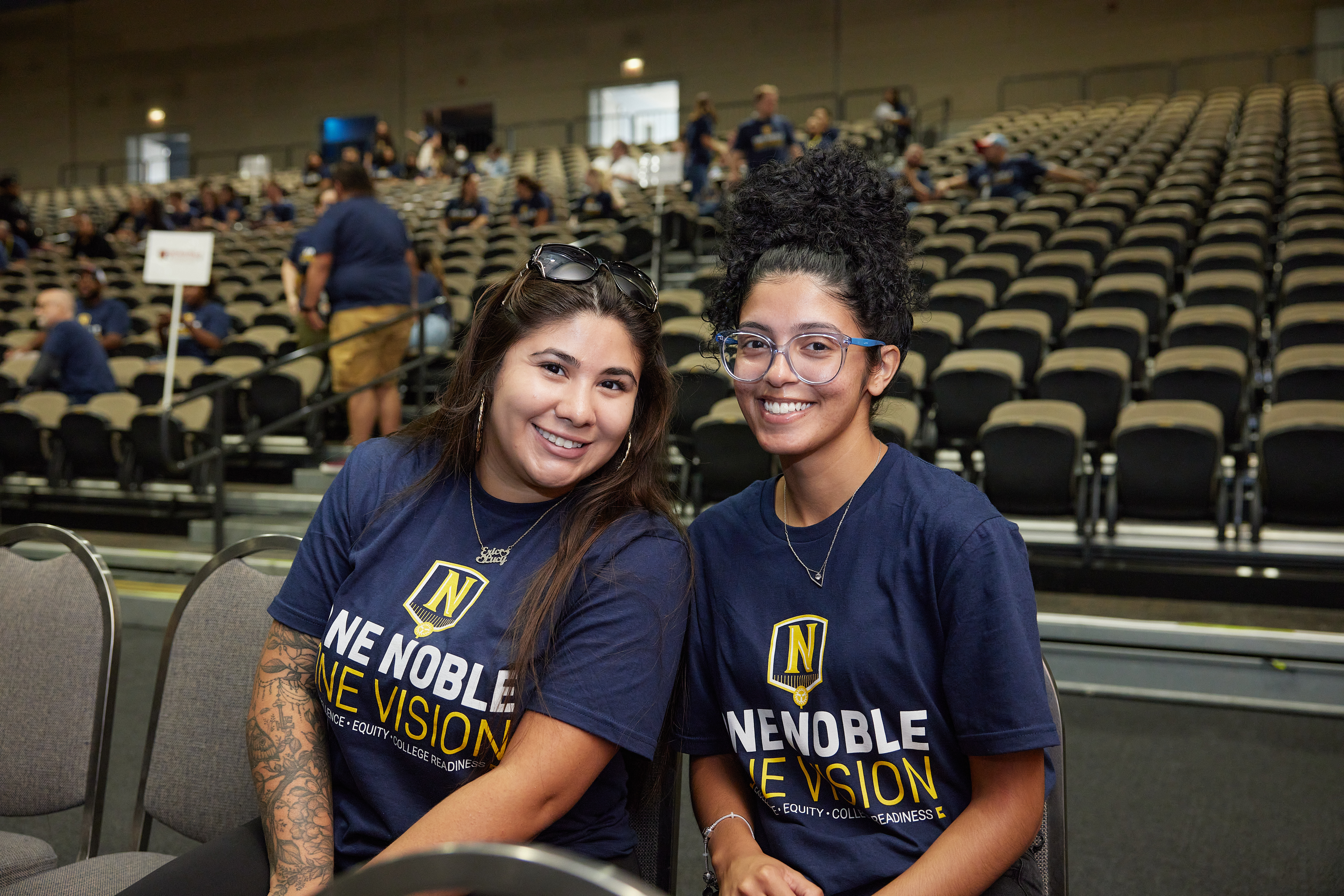 In this photo, two Noble Schools' staff members are sitting and smiling at the camera. They are in an auditorium with hundreds of chairs behind them. They are both wearing dark blue shirts that have the Noble Schools logo on them and say "One Noble, One Vision: Excellence, Equity, and College Readiness".