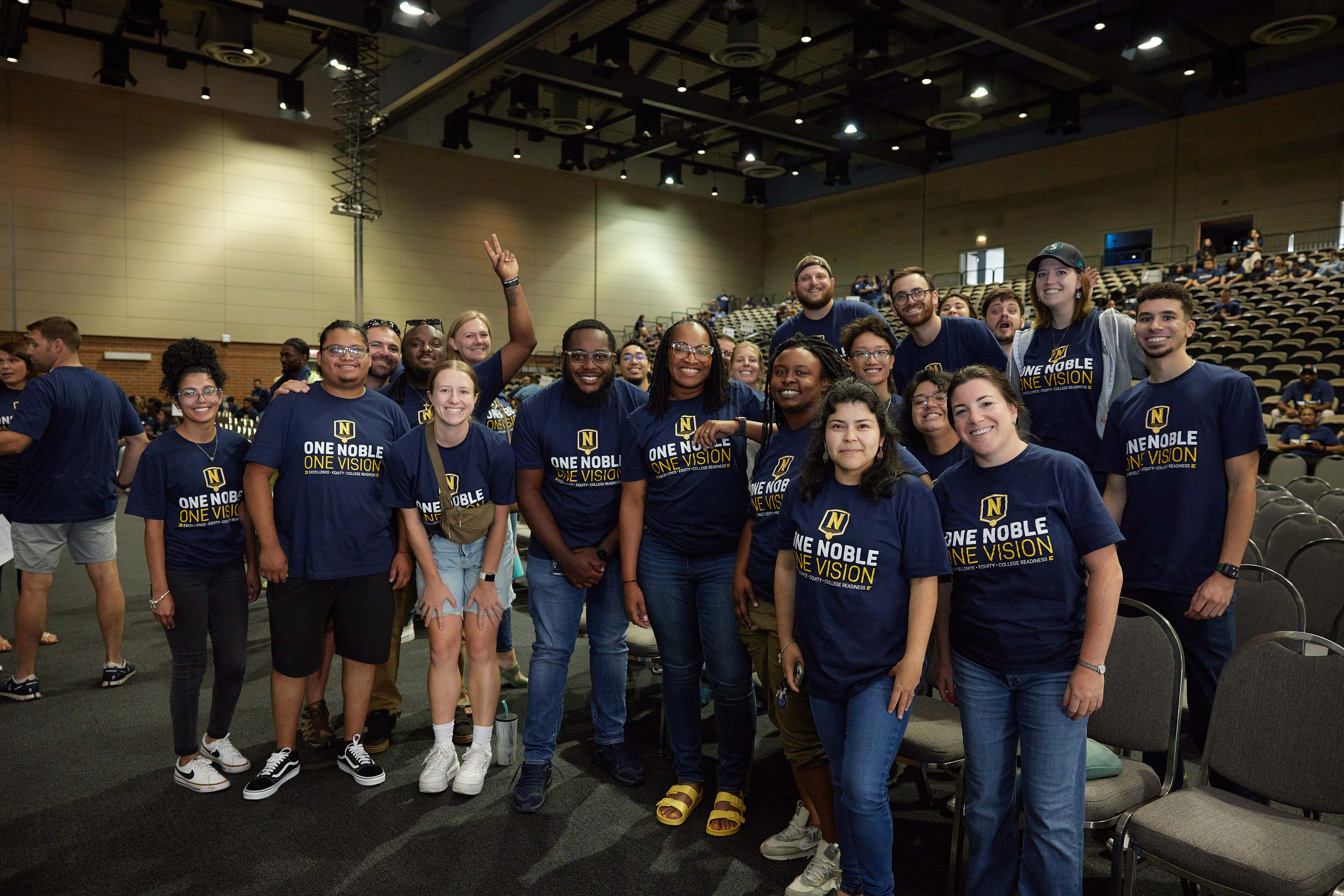 Photo shows a group shot of The Noble Academy staff standing and looking at the camera, smiling. They are in a big auditorium at UIC with hundreds of chairs behind them and bright lights above them. They are all wearing dark blue shirts that have the Noble Schools logo on them and say "One Noble, One Vision: Excellence, Equity, and College Readiness".