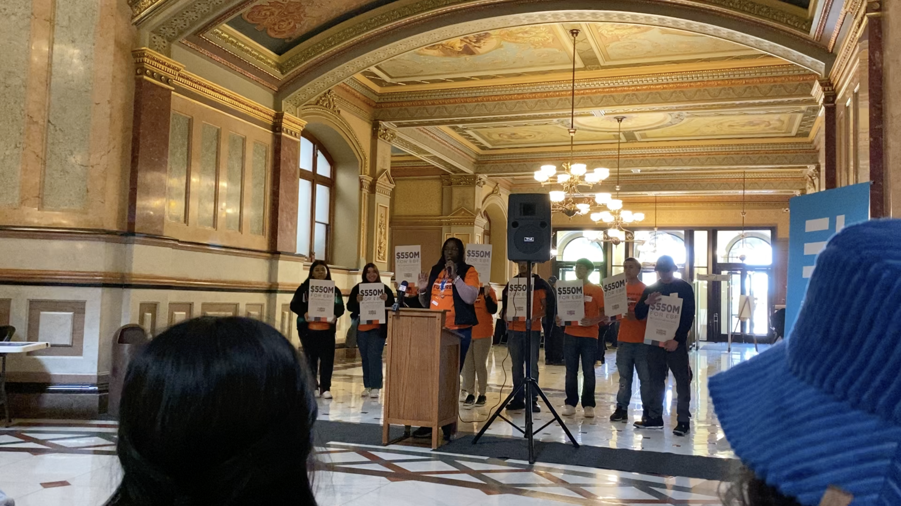 Image shows Temeka Cartwright, a parent at Butler College Prep, speaking behind a podium to a crowd in the Illinois State Capitol building. She is wearing a bright orange shirt that reads "$550M for Evidence-Based Funding" and has the Funding Illinois Future logo on it. She is holding a microphone and has a group of other advocates behind her, wearing the same shirt and holding signs that say the same thing as the shirts. In the foreground of the image, you can see a couple of heads of audience members. This photo was taken during Noble Schools' Lobby Day trip in April 2023.