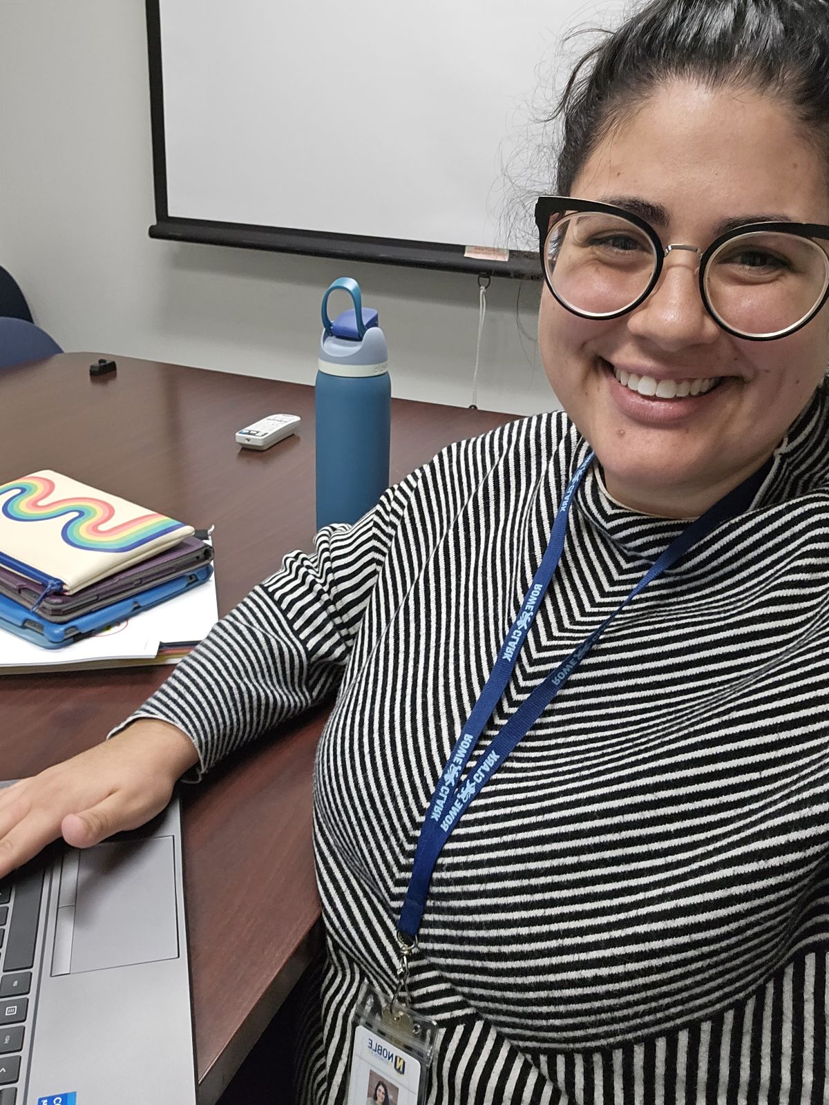 Photo shows a waist up selfie of Dr. Maria Vlantis, a school psychologist at Noble Schools. She is sitting at a desk with her laptop in front of her and is looking and smiling at the camera.