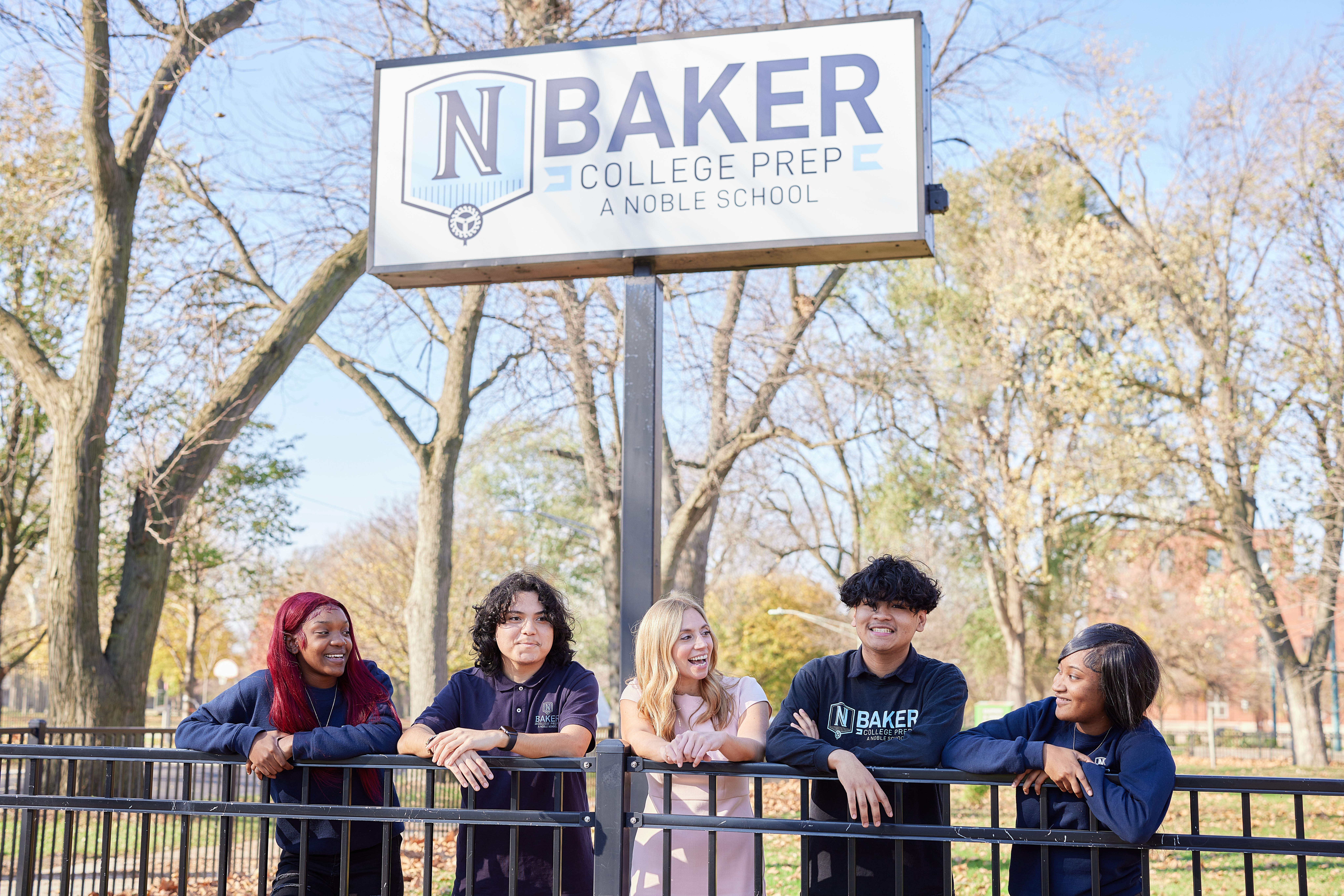 Photo shows four Baker College Prep students and their principal Mary Arrigo standing outside beneath the Baker College Prep school sign. They are smiling and look like they are talking to each other. They lean against a black fence in front of them.