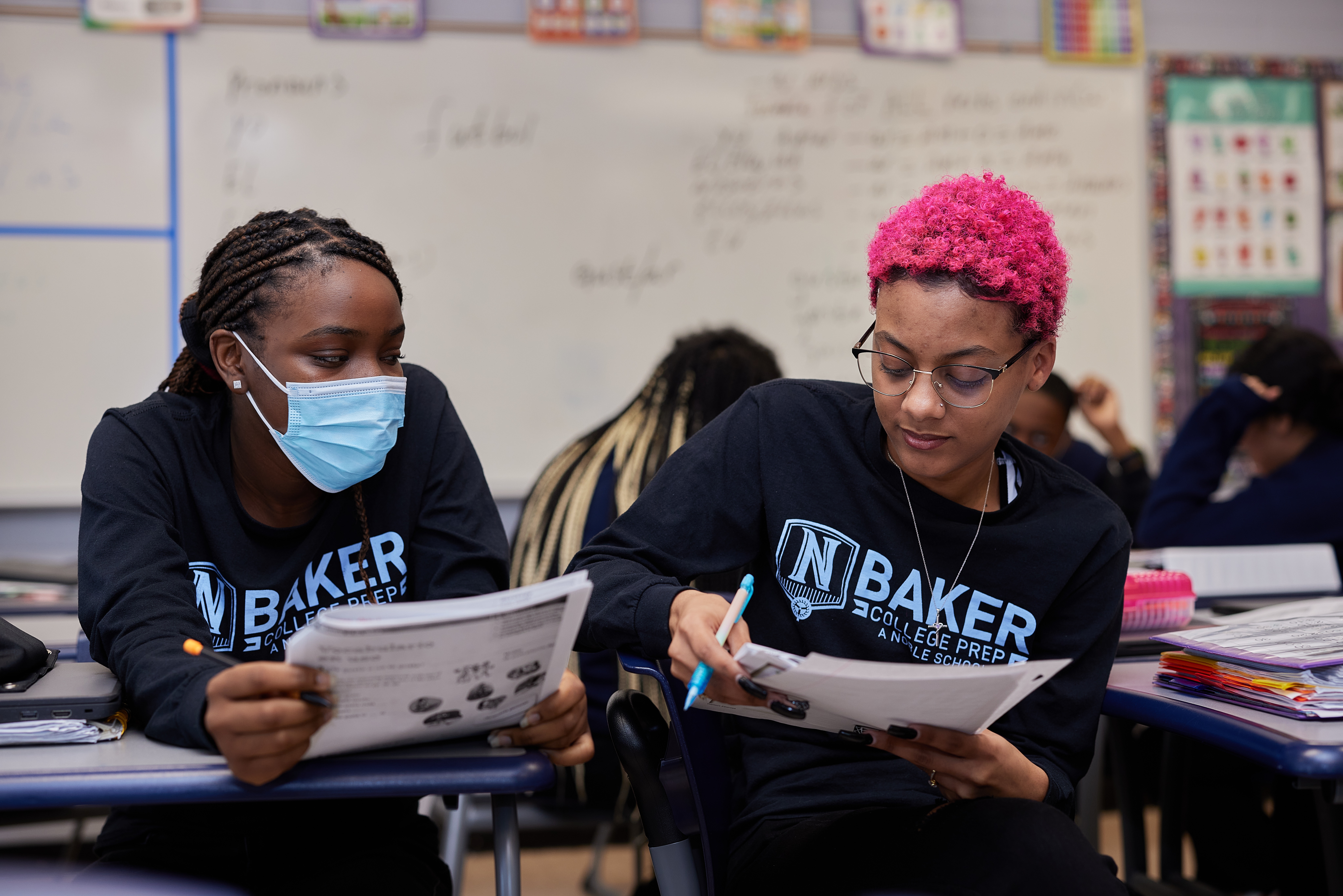 Photo shows two Baker College Prep students studying together in a classroom. They are holding and looking at activity packets in their hands. They are two young Black students, one with bright pink short hair and the other with long black braids. They both wear matching Baker College Prep sweatshirts.