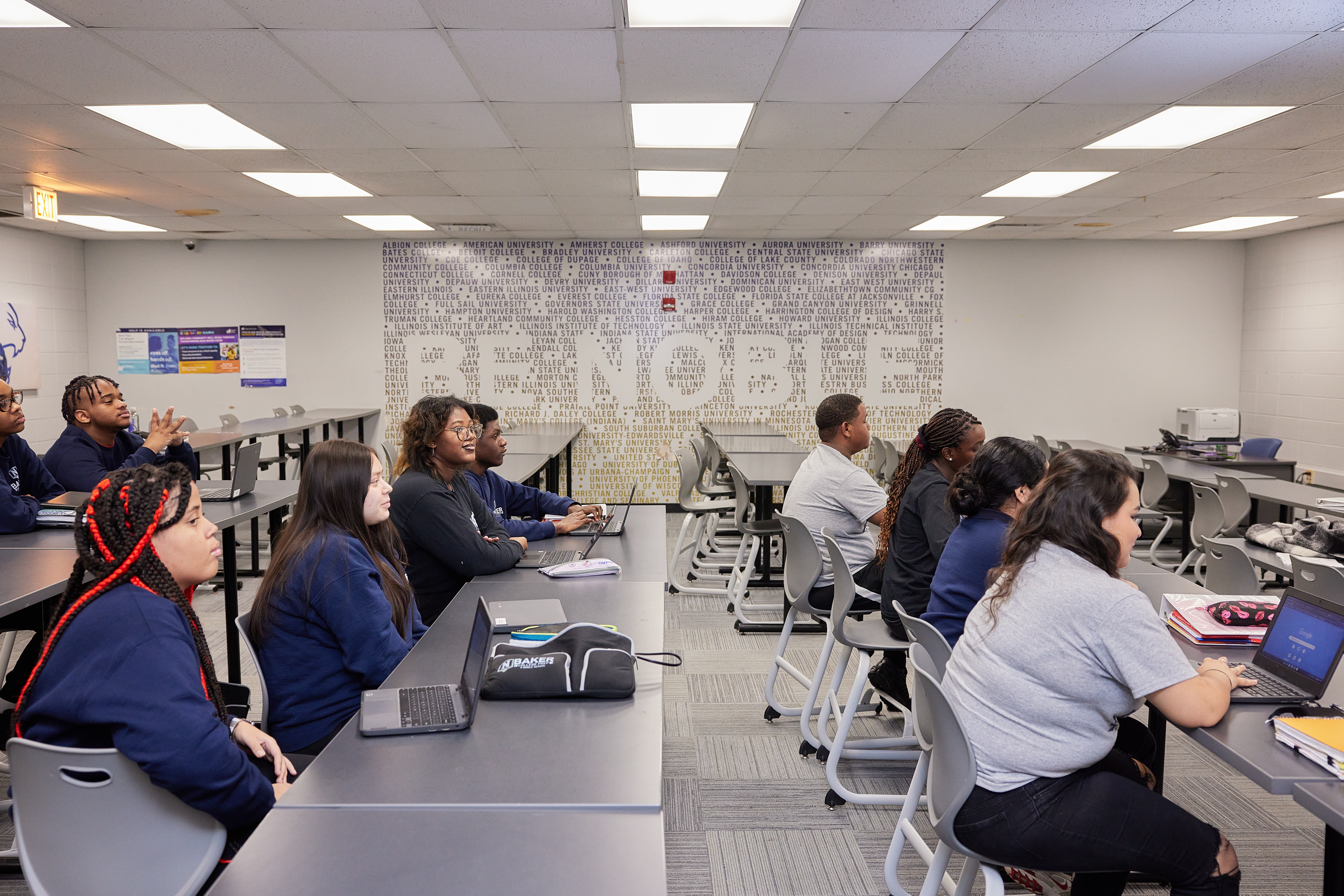Photo shows a wide shot of a Noble Schools' classroom. Black and Brown students sit behind rows of long tables and look towards the front of the classroom, which is to the right. Behind them is a wall that is covered in sticker decals that list of names of several universities and colleges. In the middle of the stickers, there is white space that is cut out to read "Be Noble" in the midst of all the university names.