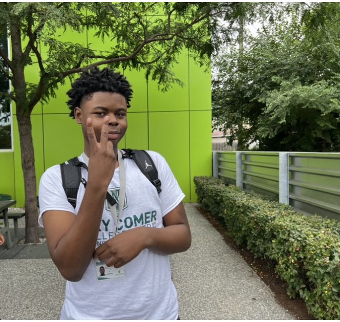 Photo shows Davontae, a Gary Comer College Prep high school student, posing with the peace sign outside of the high school building. He is a young Black man with short dark brown hair. He is wearing a gray Gary Comer t-shirt and a black backpack.