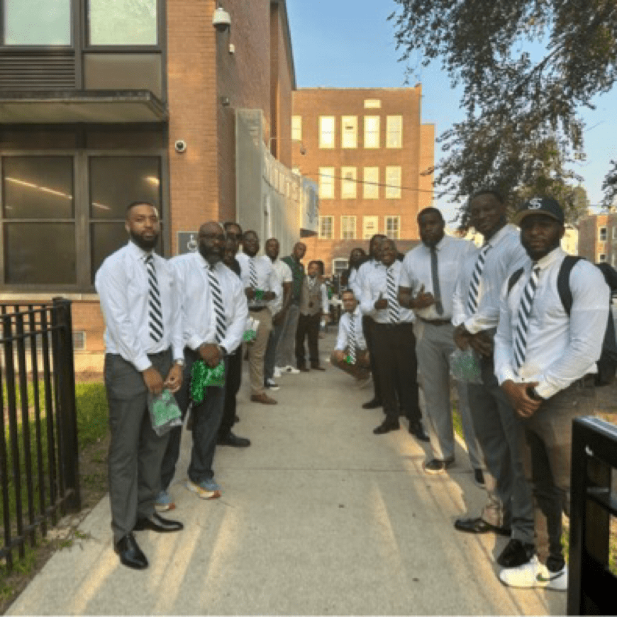 Photo shows Black male staff at Gary Comer College Prep standing in two lines on both sides of the sidewalk that leads to the front door of the school building. They are smiling at the camera. All of them are dressed in business slacks and button-up white shirts. Most of them have green and white ties, the colors of the school.