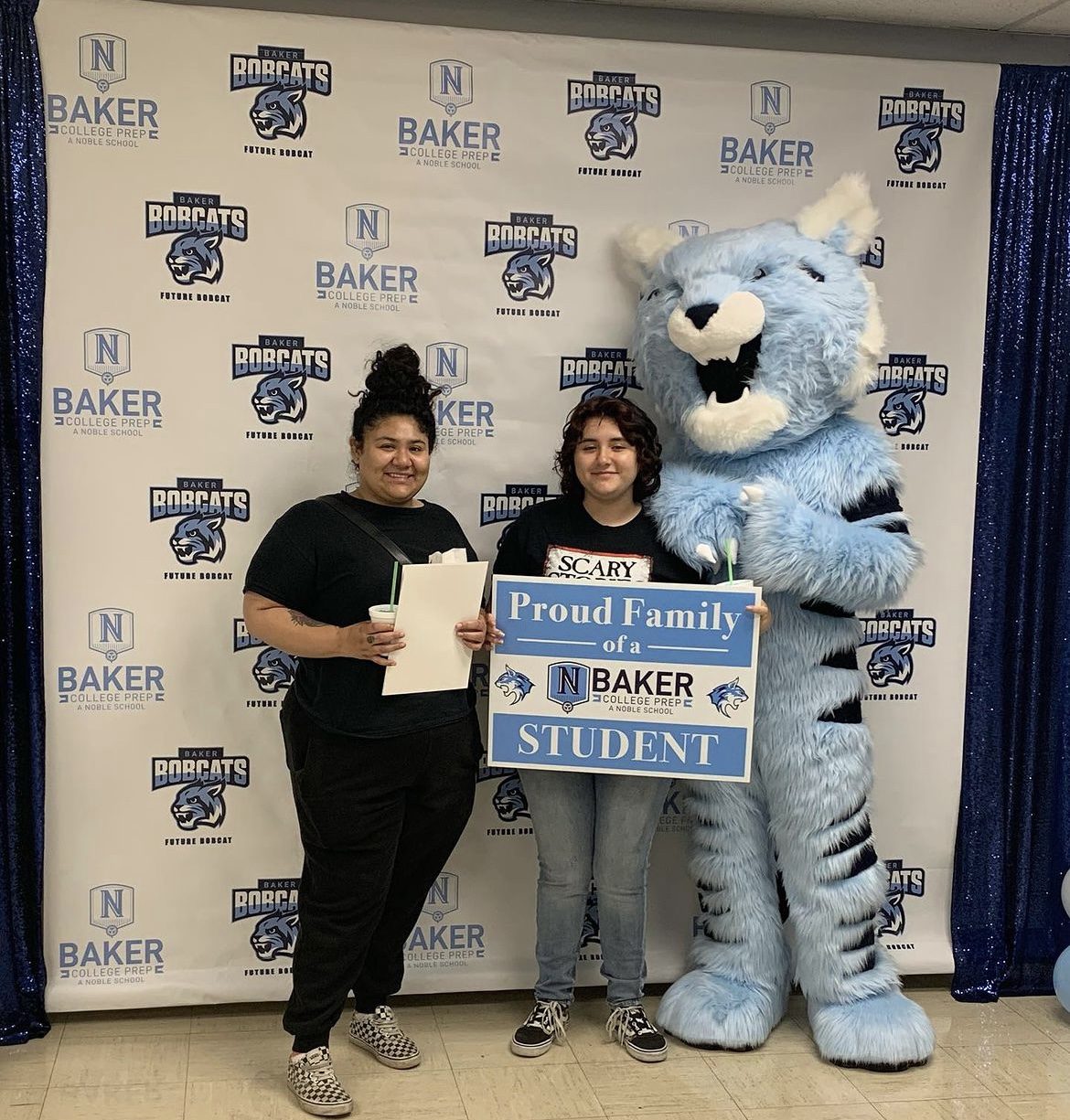 Photo shows a new Baker College Prep student and one of their family members posing with the Baker Bobcat mascot in front of a backdrop with the Baker logo. The student holds a light blue and white sign that has the Baker logo on it and says "Proud Family of a Baker College Prep Student"
