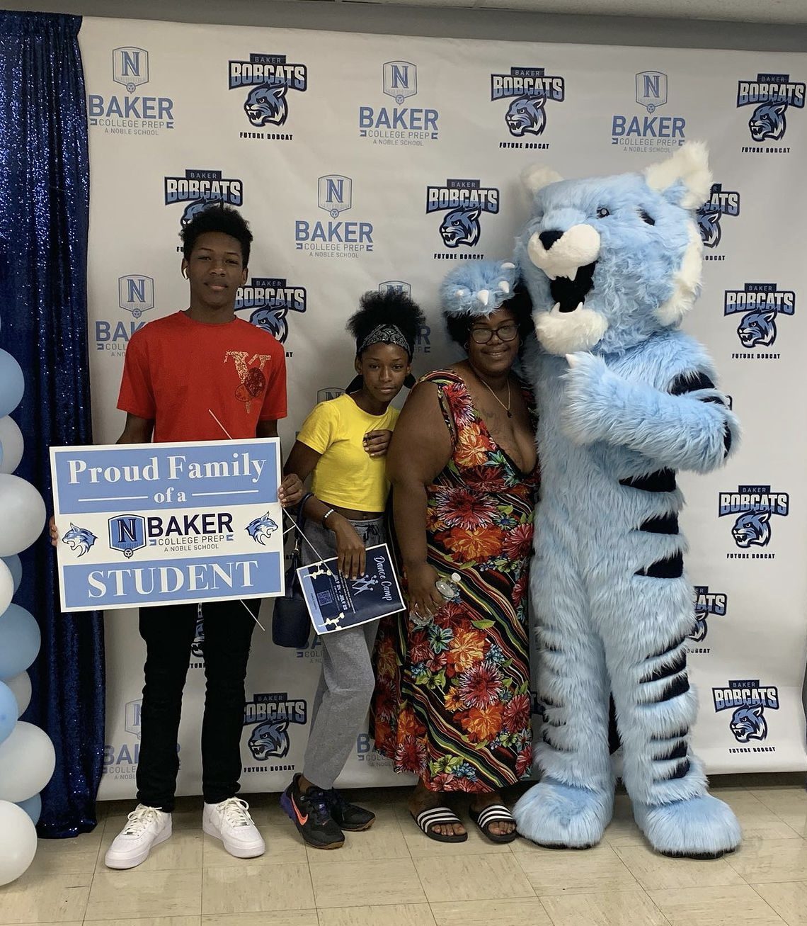 Photo shows a new Baker College Prep student and their two family members posing with the Baker Bobcat mascot in front of a backdrop with the Baker logo. The student holds a light blue and white sign that has the Baker logo on it and says "Proud Family of a Baker College Prep Student"