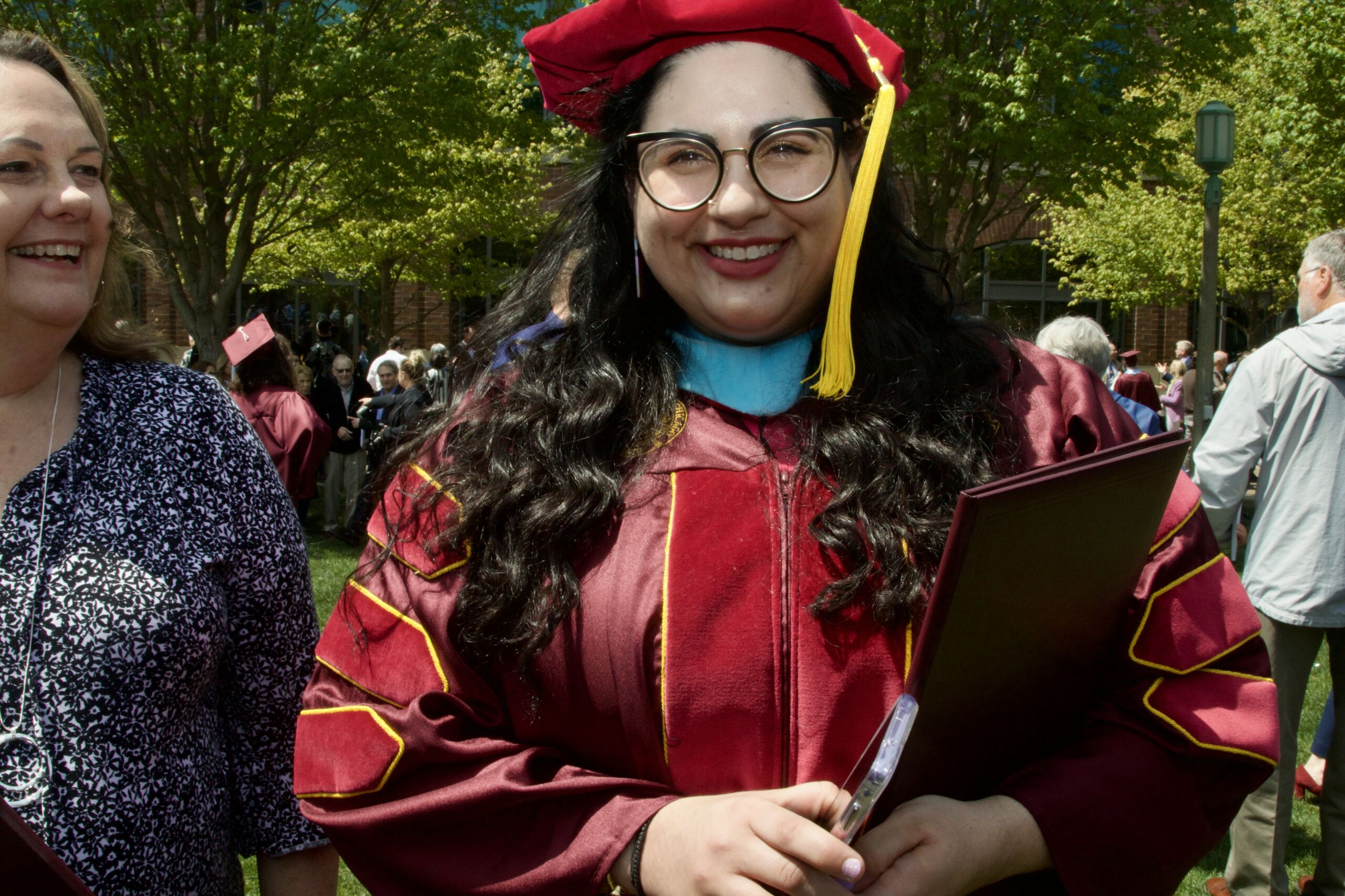 Photo shows a shoulders up shot of Dr. Maria Vlantis, school psychologist at Noble Schools, in red Loyola University graduation robes. She is smiling at the camera and is holding her diploma in one hand and her phone in the other. Around her are several other people and graduates taking photos in the background.