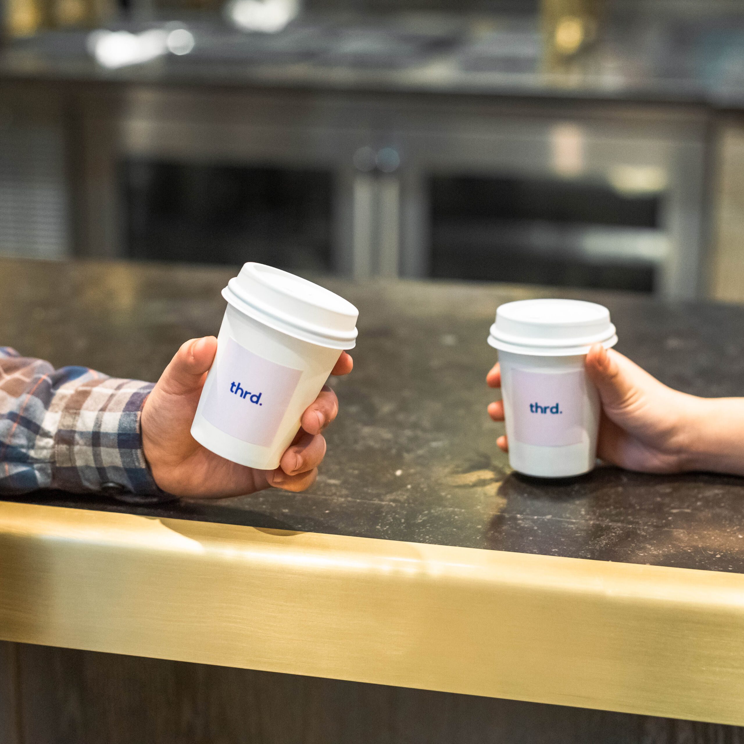 Photo shows two hands holding Thrd Coffee Company branded coffee cups. The hands are leaning against a countertop.