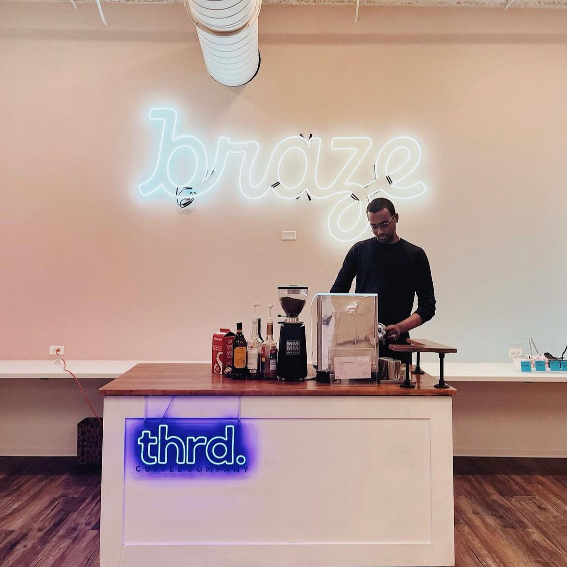 Photo shows the Thrd Coffee Company mobile coffee cart inside an office space. An employee stands behind the cart, pouring some coffee. Behind him and the cart is a wall that has a white neon sign on it that says "brave" in a script font.