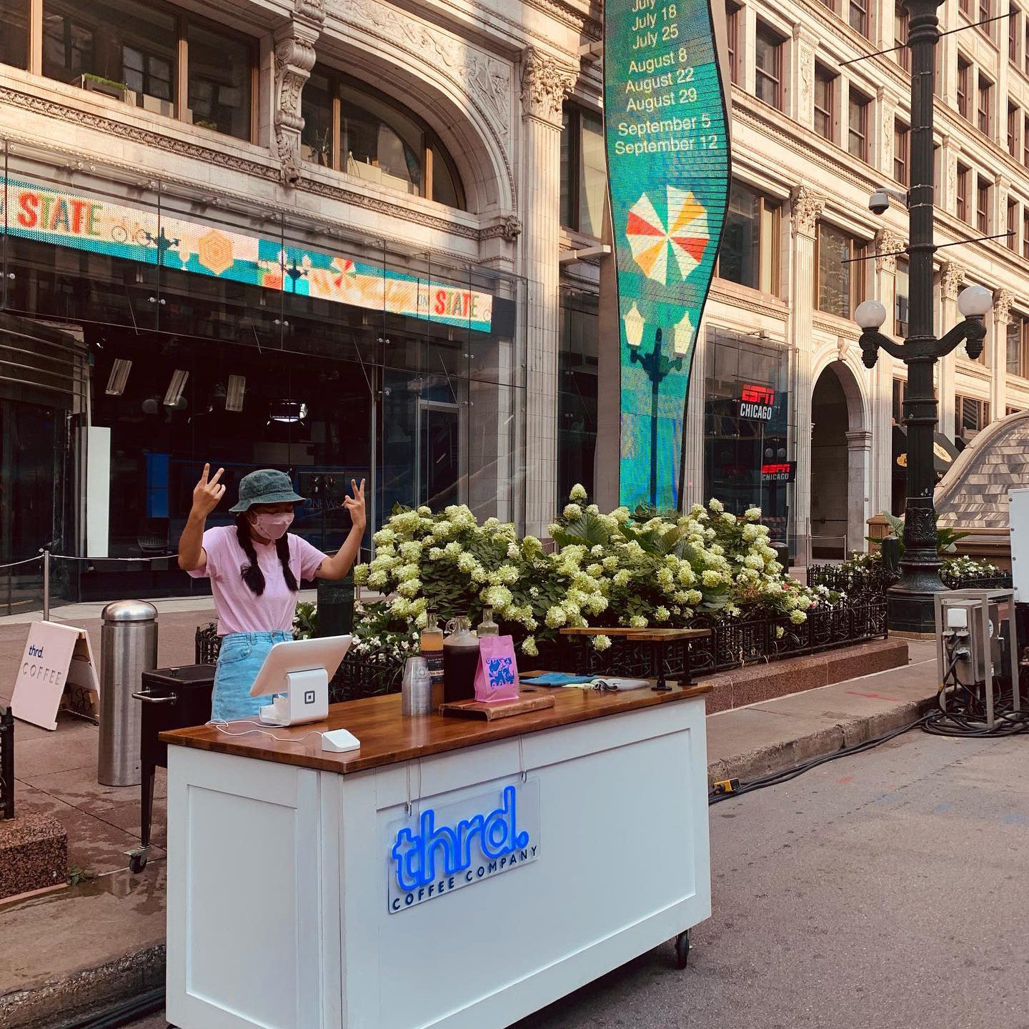 Photo shows the Thrd Coffee Company mobile coffee cart outside in downtown Chicago. The cart looks like a simple white wood counter with a brown wood top. An employee stands behind the cart, posing at the camera with peace signs.