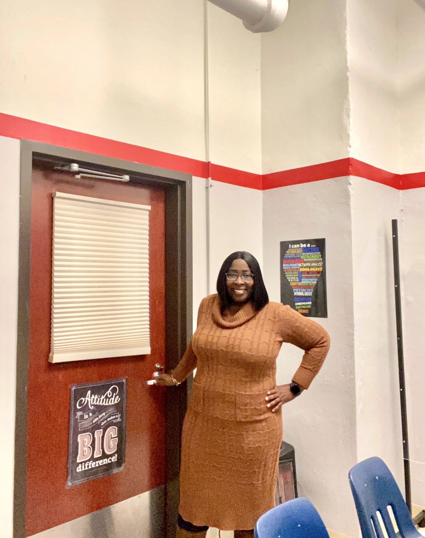 Photo shows a full body shot of Dr. Tiffani Farrow, a school psychologist at Noble Schools, as she stands in front of her office door at UIC College Prep, holding the handle of the door as she is about to go in. She is looking and smiling at the camera with her other hand on her hip.