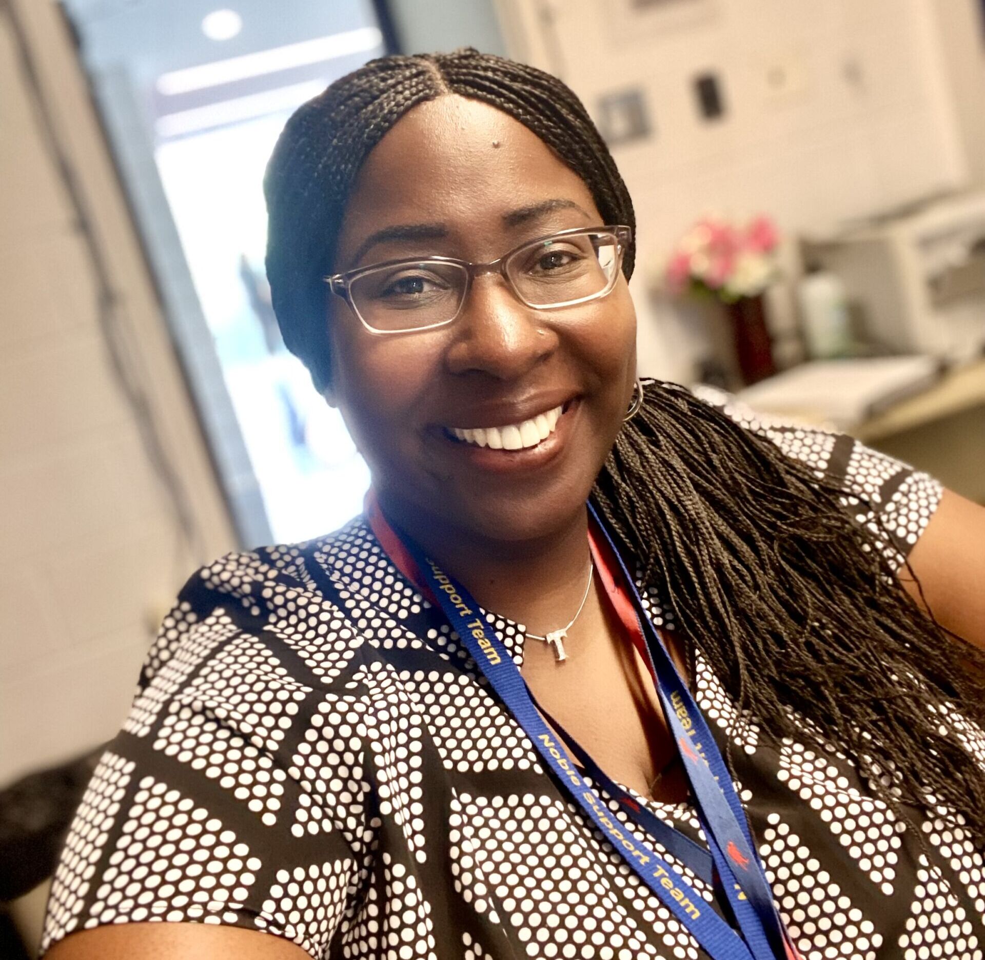 Photo shows a shoulders and up selfie of Dr. Tiffani Farrow, a school psychologist at Noble Schools. She is wearing a dark blue lanyard that has yellow text on it that reads "Noble Support Team". In the background, you can see some of her office at Johnson College Prep.