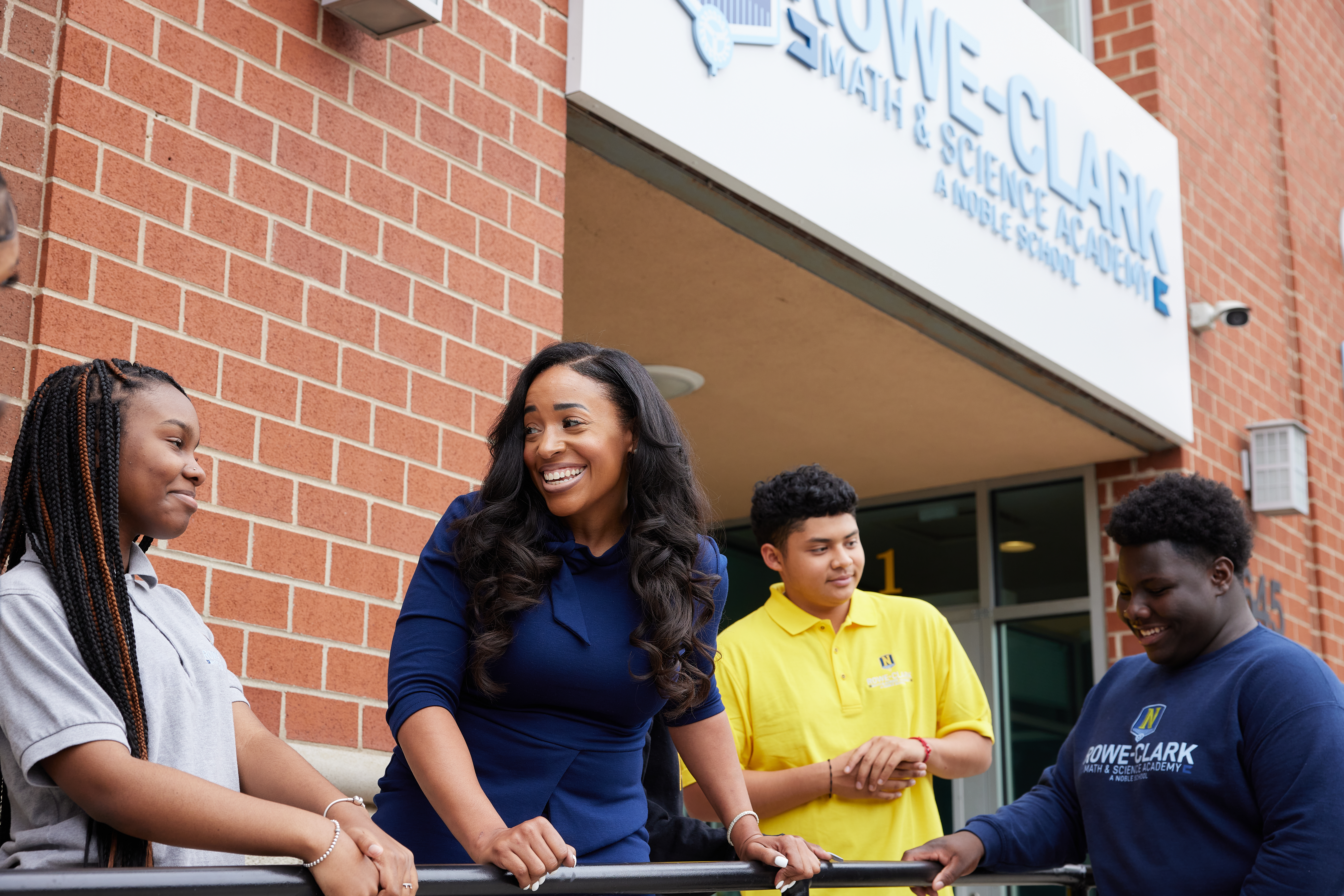 Photo shows Principal Moss talking with three students outside the front entrance of Rowe-Clark Math and Science Academy. They are all smiling and looking at each other like they are in a good conversation. You can see the Rowe-Clark sign in the background.