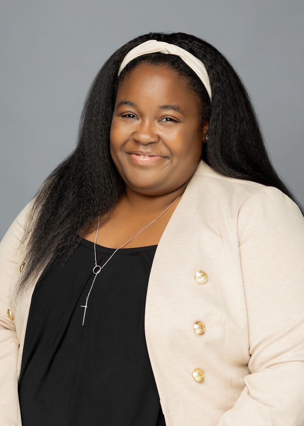This photo is a professional headshot of Brittany Miller. She is a Black woman with long, straight black hair. In this photo, she is wearing a cream white headband that matches her cream white blazer. She is wearing a black blouse underneath the blazer and has a simple silver necklace and earrings on. She is smiling and her arms are relaxed to her sides with her hands slightly crossing in front.