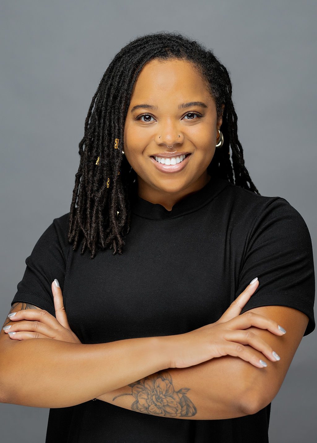 This photo is a professional headshot of Brittany Parks. She is a Black woman with medium-length black locs. She is wearing a simple black blouse, gold earrings, and has a nose ring, two nose studs, and a few gold loc jewelry pieces in her hair. She also has an intricate all-black tattoo of a flower on her left forearm. She is smiling and crossing her arms confidently.