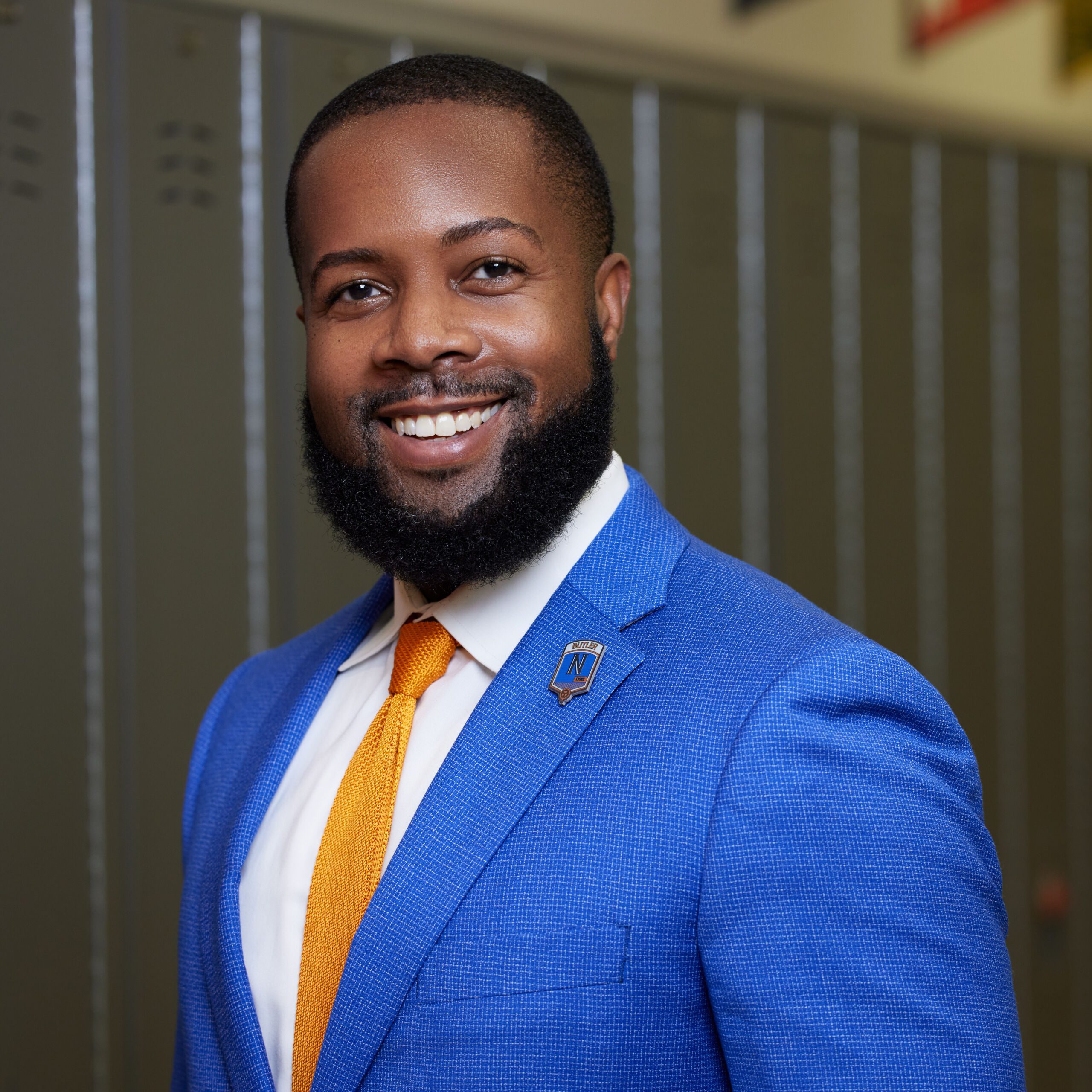 Photo shows a professional headshot of Brian Riddick, the principal of Butler College Prep. He is a Black man with short black hair and a short beard. He is wearing a bright blue suit with a white button up and a bright orange tie, the colors of Butler College Prep. He is smiling and looking at the camera. You can see lockers at Butler behind him.